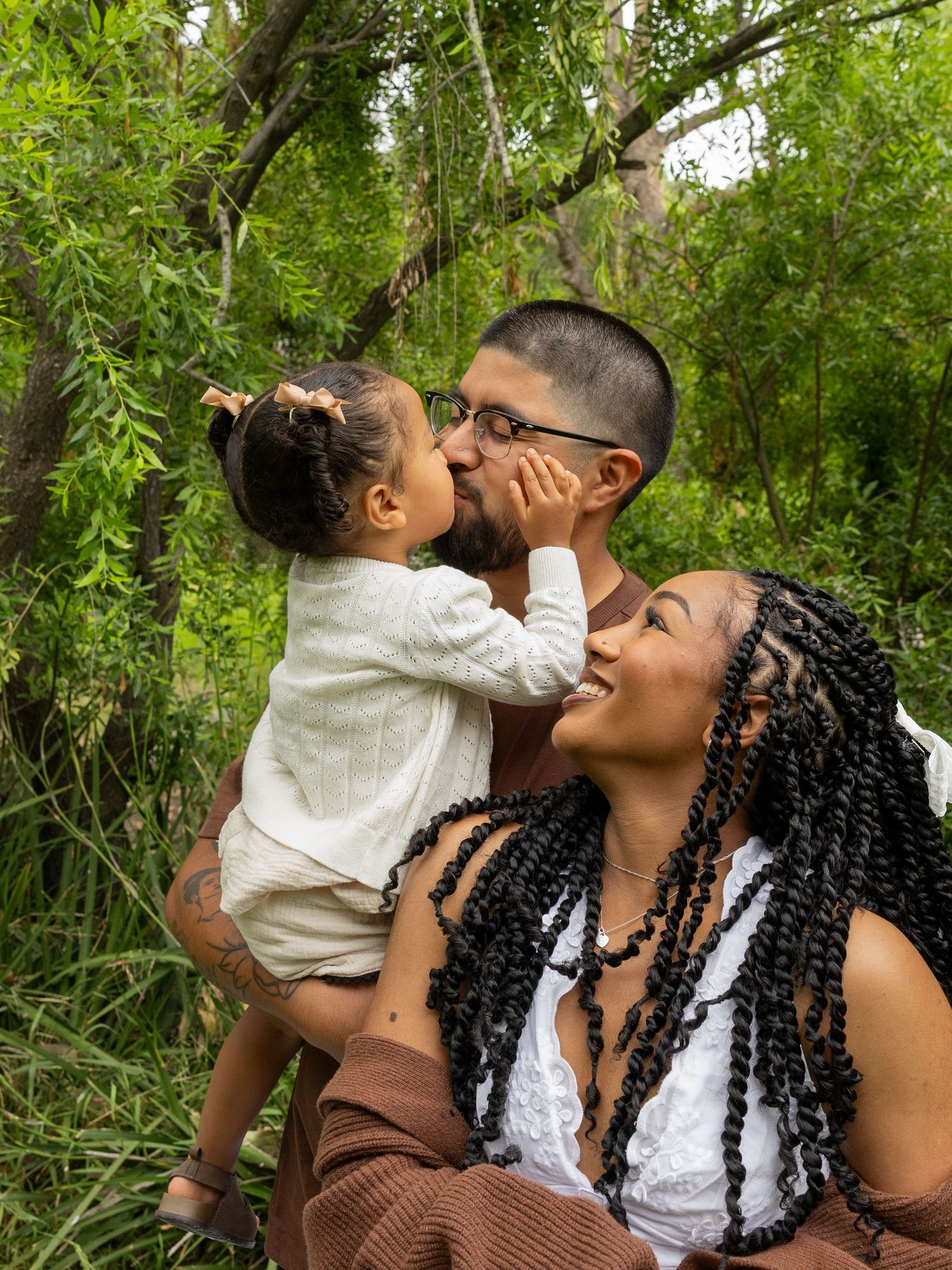 A man holds a toddler who kisses his cheek while a woman with braids smiles joyfully outdoors.