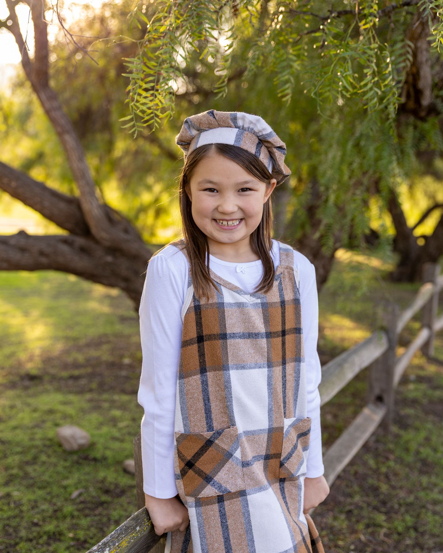 Young girl smiling outdoors in plaid pinafore dress and beret, standing by wooden fence with green trees.