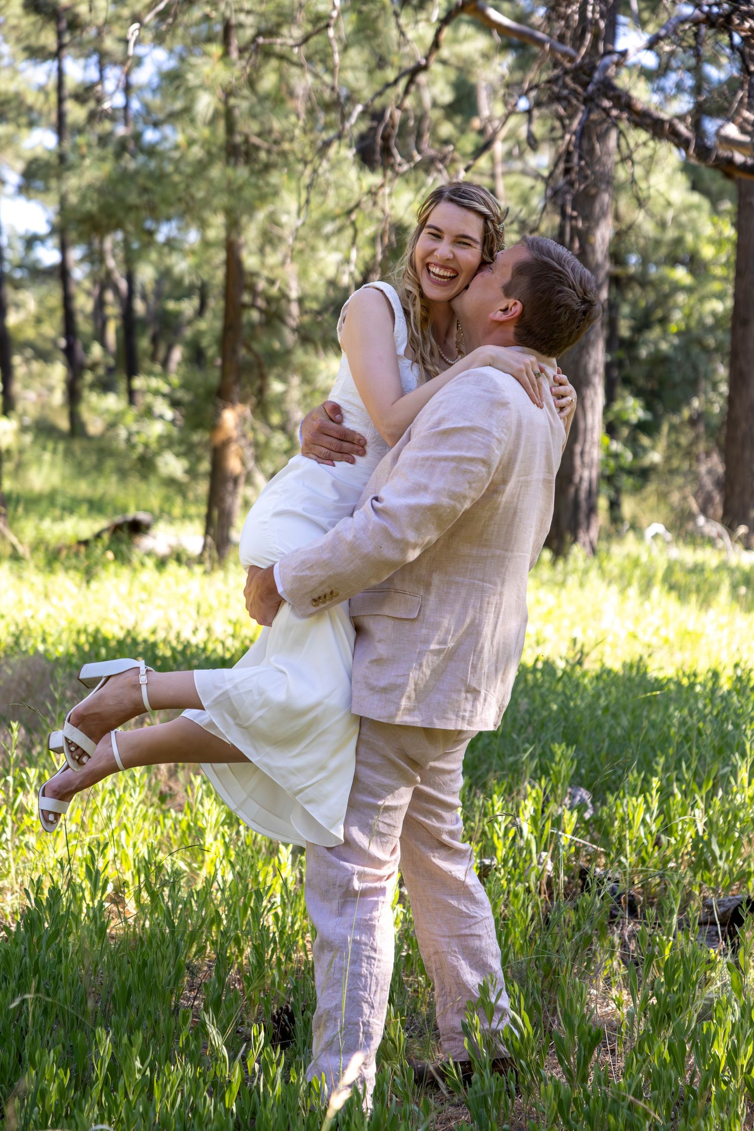 Groom lifting bride in white dress, laughing joyfully in a sunlit forest clearing during outdoor wedding.