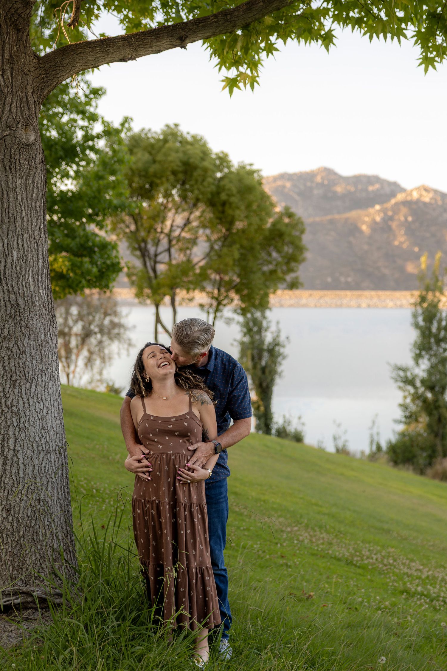 Pregnant couple embracing under a tree by a lake, man kissing woman's cheek while she smiles, mountains in background.