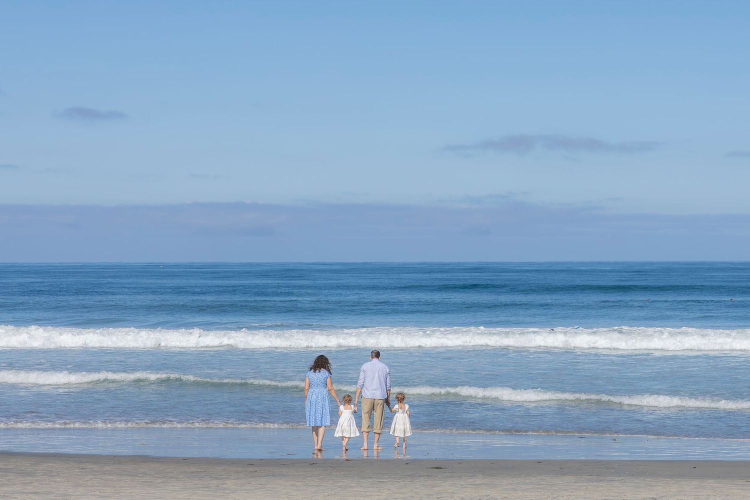 Family of four holding hands walking toward ocean waves on a sunny beach with blue sky.