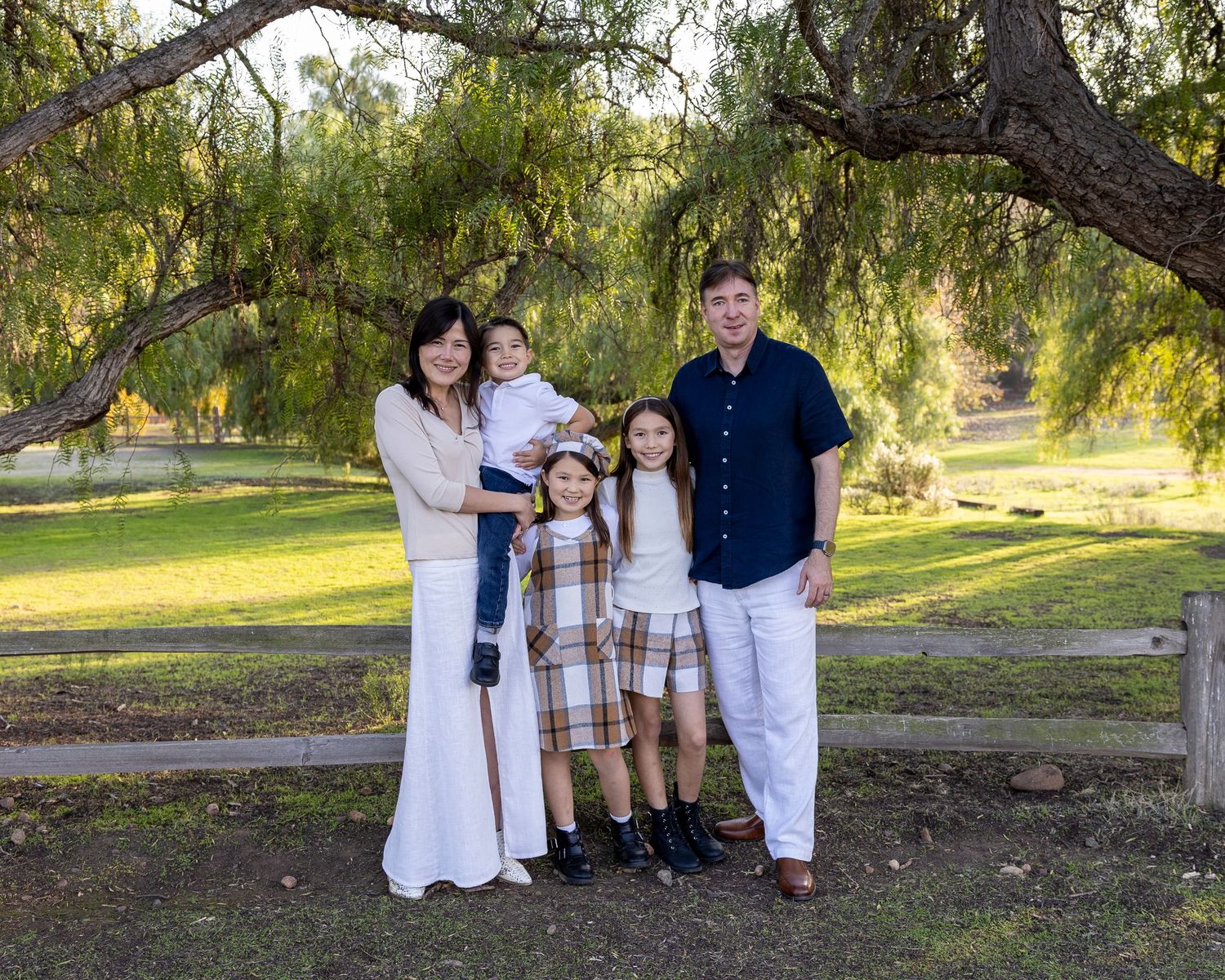 Family of five posing outdoors under a willow tree near a wooden fence in a lush green park.
