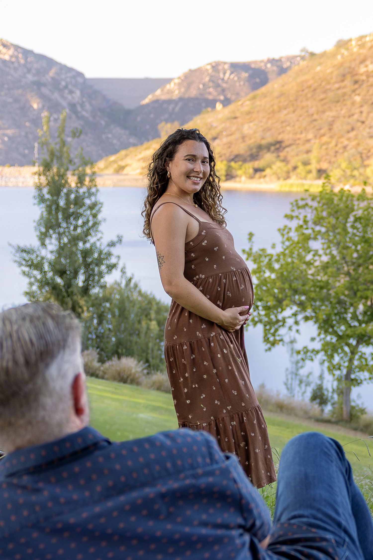 Smiling pregnant woman in a brown floral dress cradling her baby bump near a serene mountain lake. San Diego photographer
