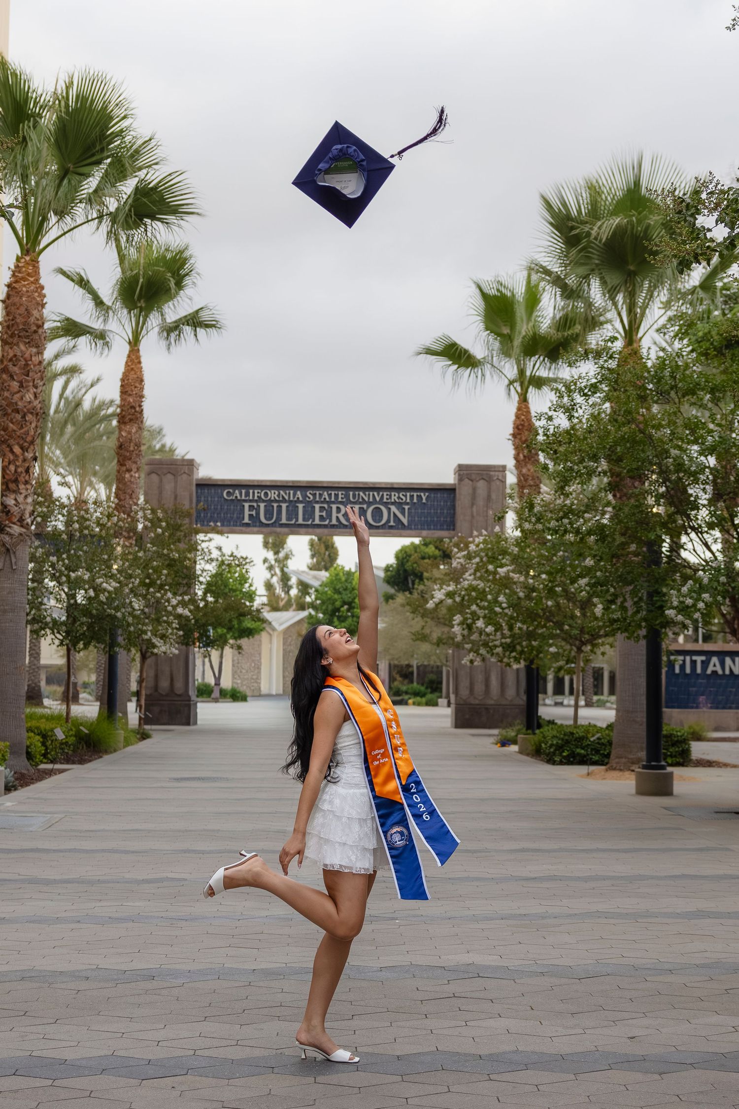 A graduate tosses her cap in the air at California State University Fullerton, wearing a white dress and honor sashes. San Diego photographe