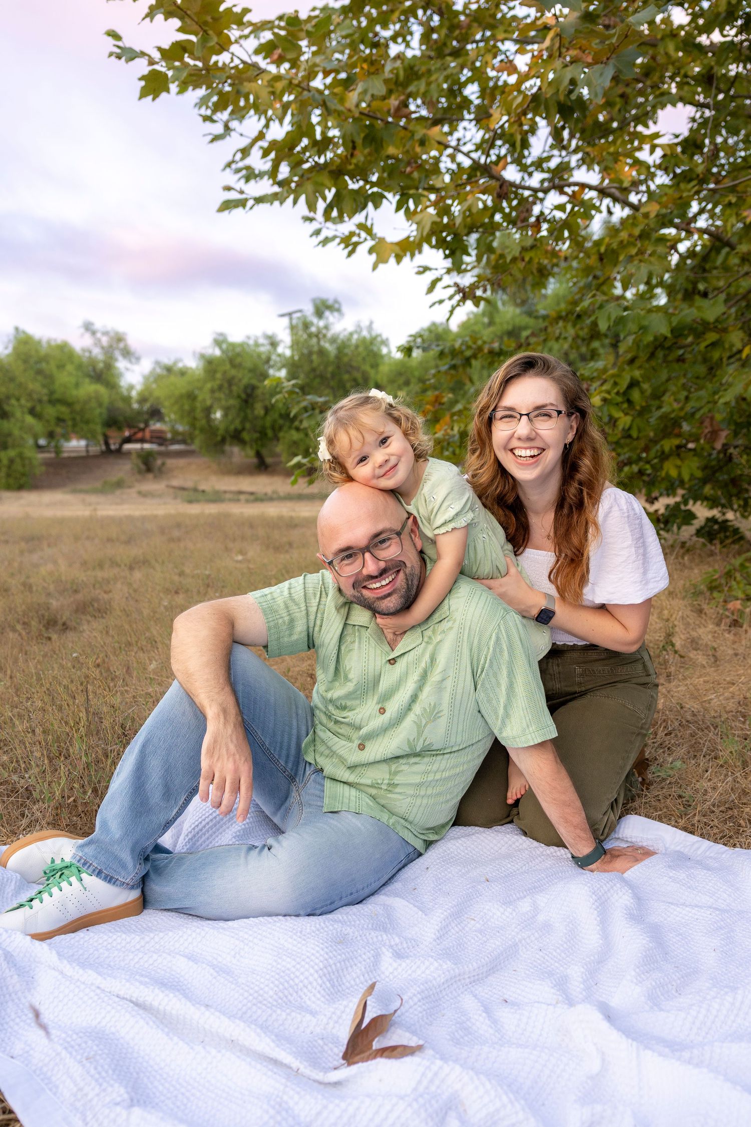 A happy family of three sits on a white blanket outdoors, smiling together beneath a leafy tree in a grassy field.