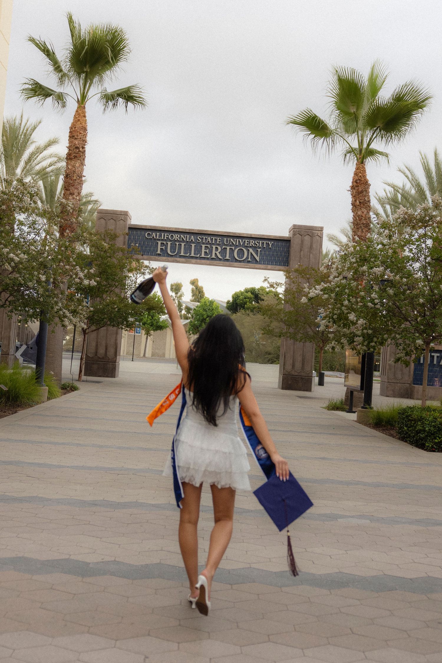 Graduate celebrates at California State University Fullerton entrance, holding cap and diploma in white dress. San Diego photographer