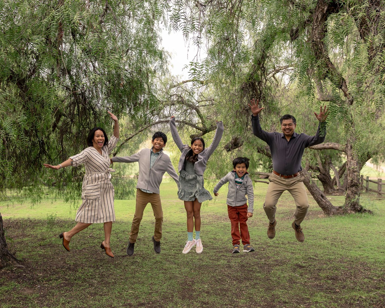 A joyful family of five jumping together outdoors under willow trees on green grass.