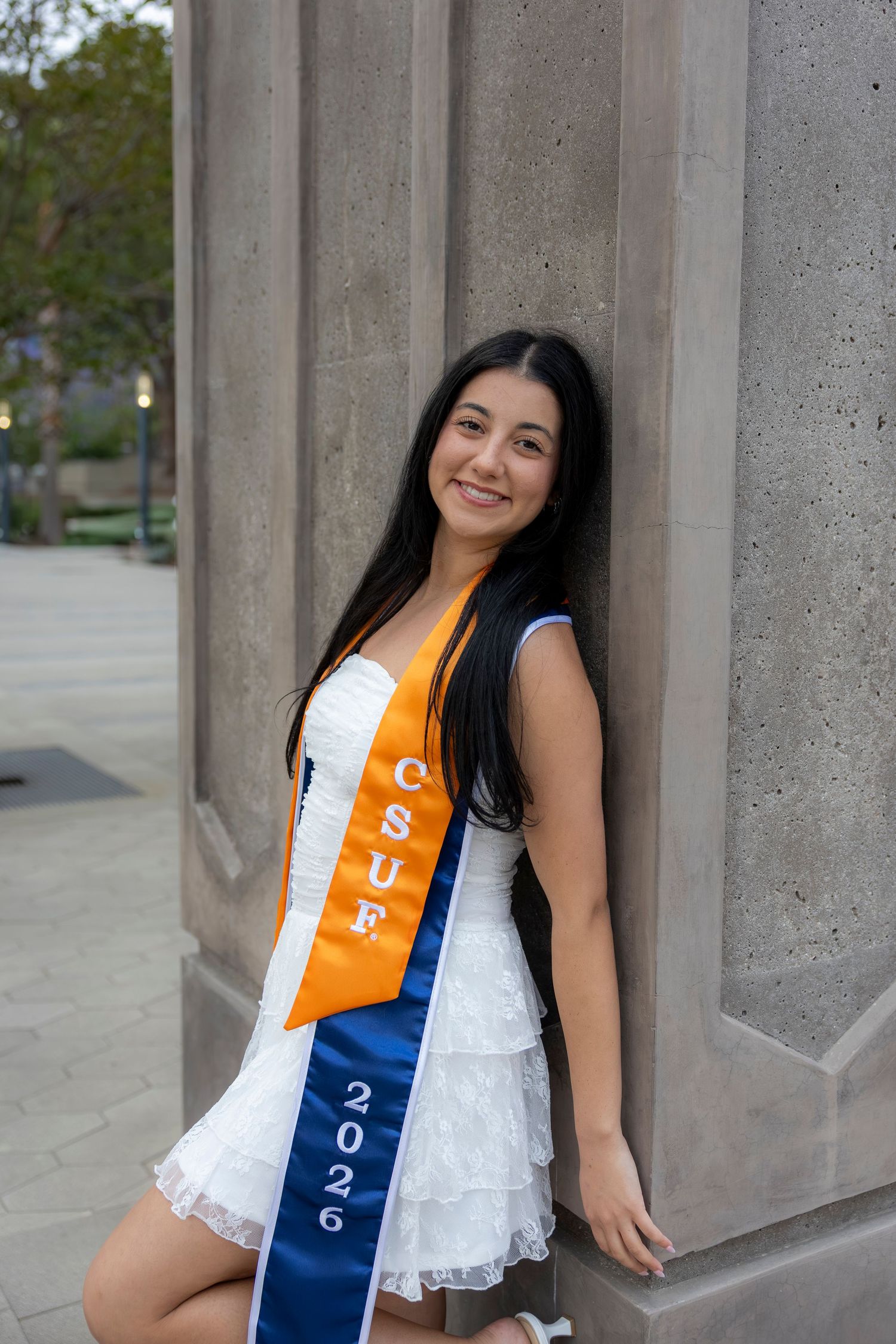 Young woman in white dress wearing CSUF graduation sashes for 2026, smiling while leaning against a stone column. San Diego photographer