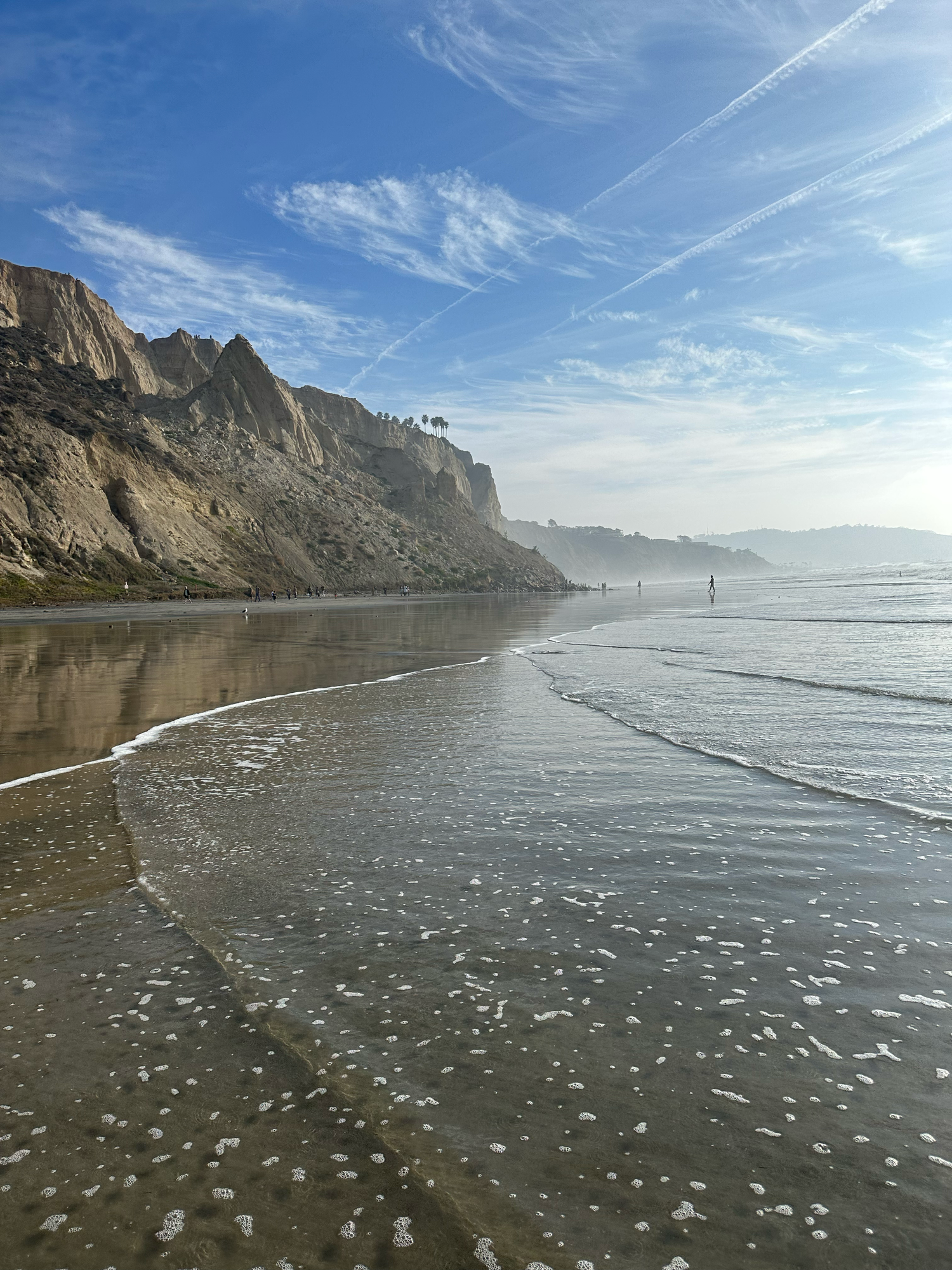 Scenic California beach with dramatic sandstone cliffs, wet reflective sand, ocean waves, and a clear blue sky with wispy clouds.