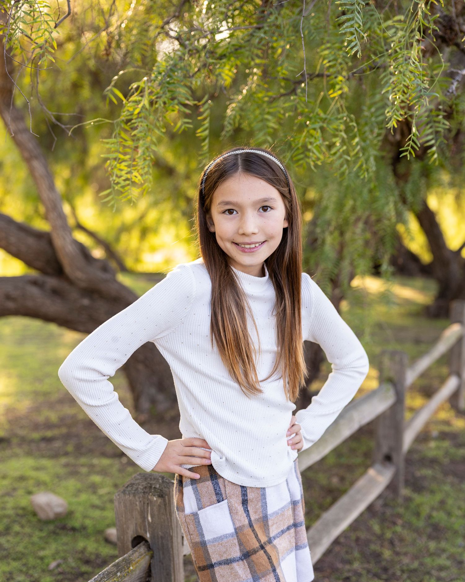 A young girl with long hair poses confidently with hands on hips near a wooden fence outdoors.