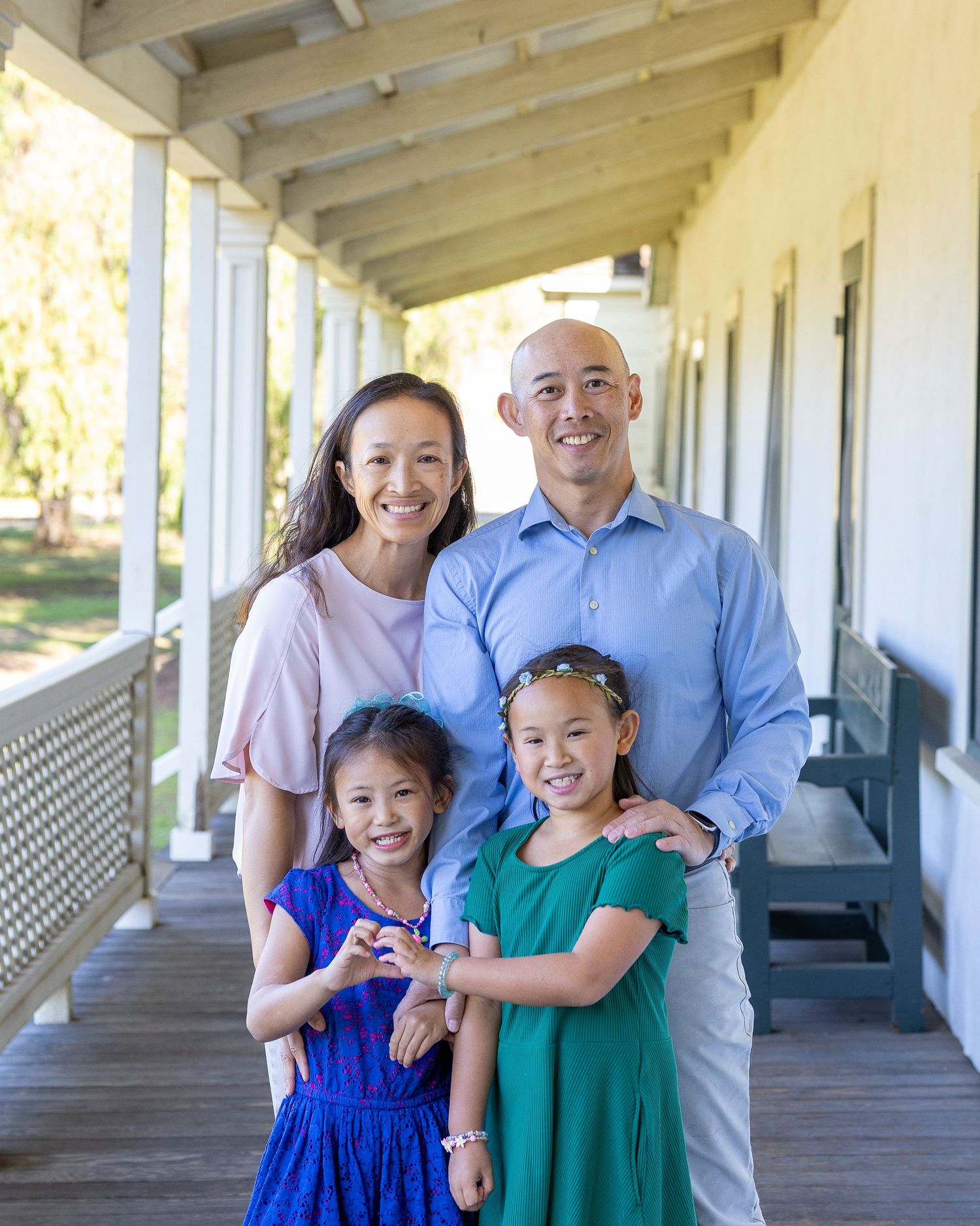 Family of four smiling on a porch, parents standing behind two young girls in blue and green dresses.