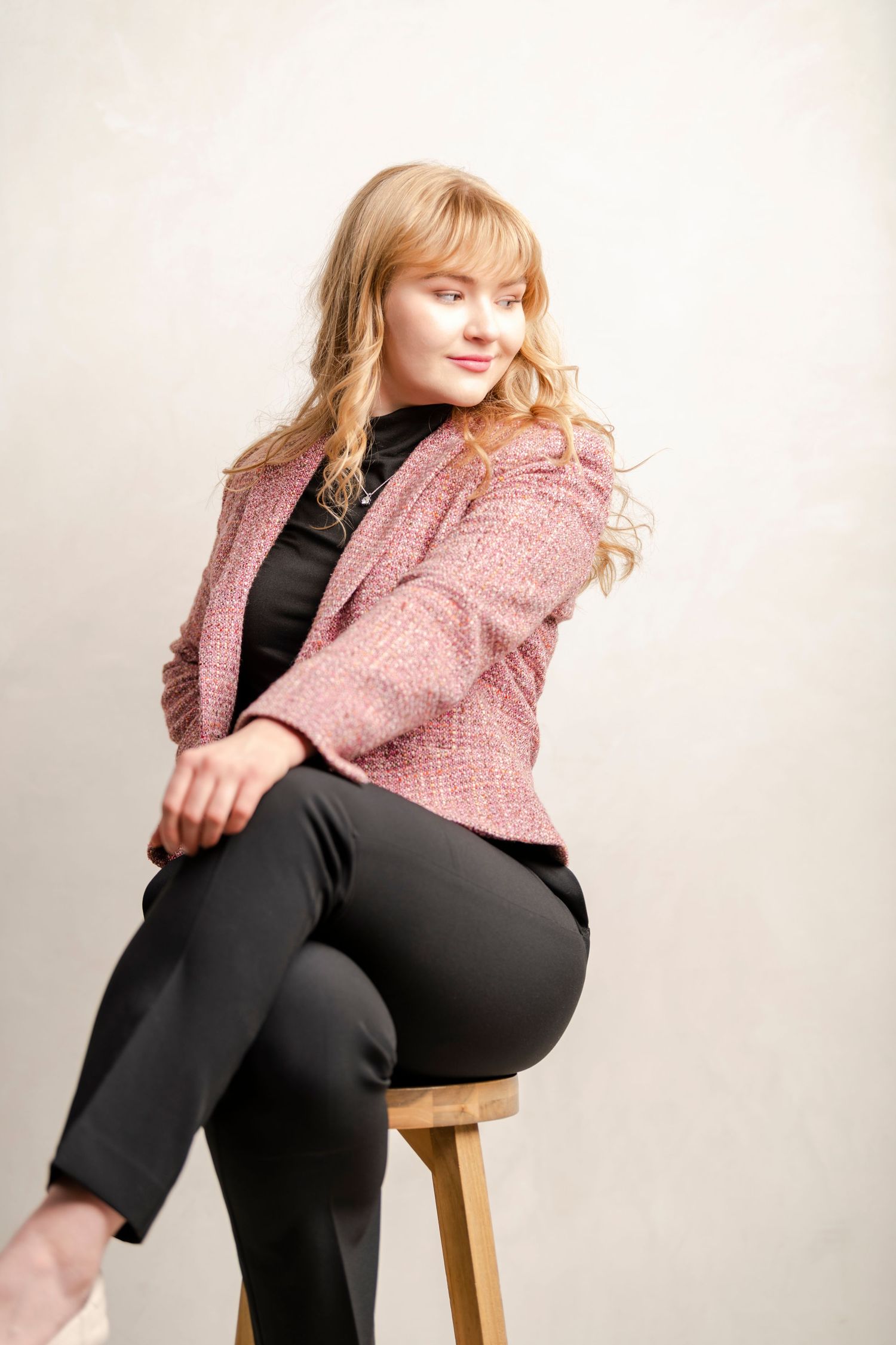 Studio portrait of young woman sitting on stool in blazer with soft light and clean neutral backdrop