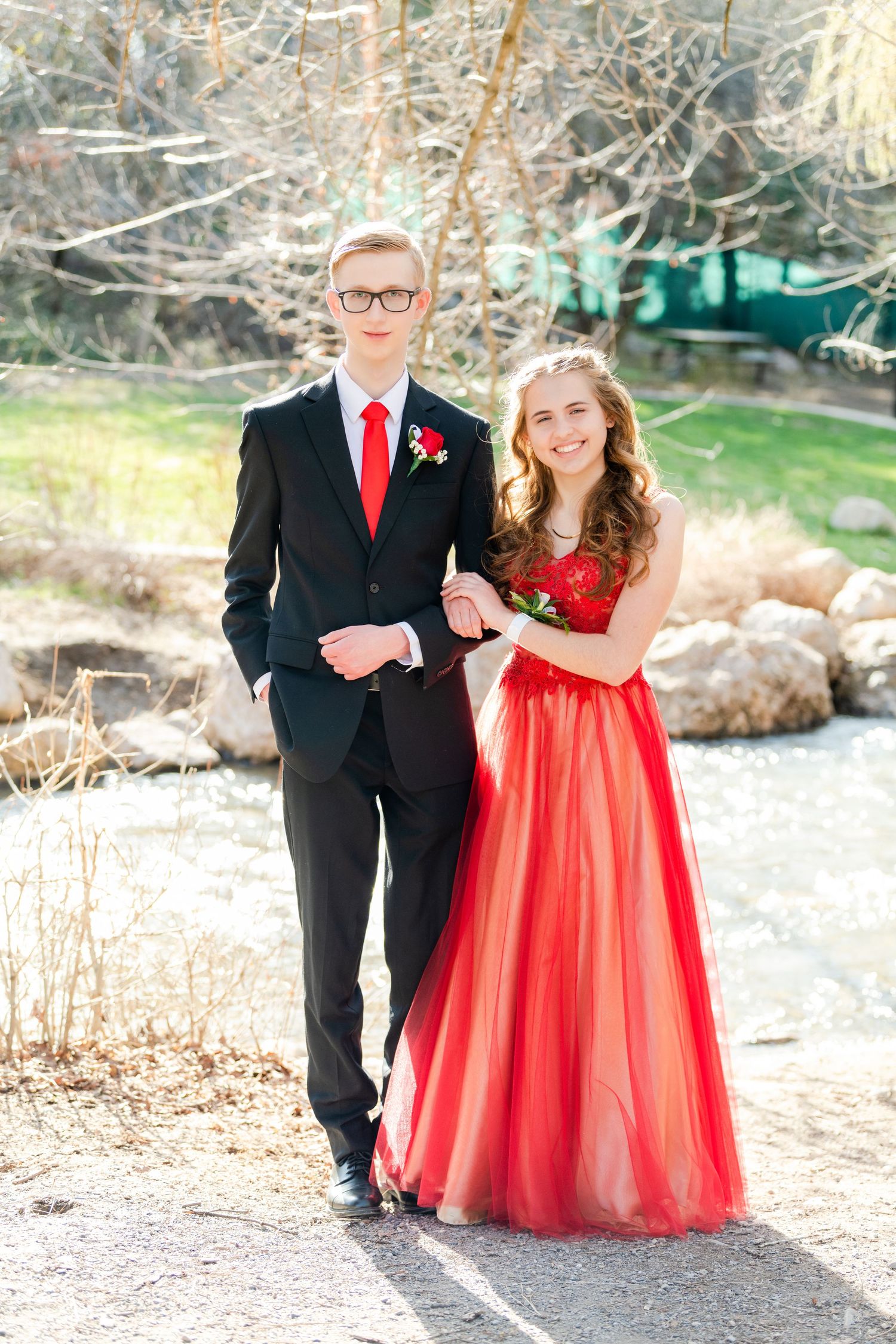 Senior Woods Cross prom couple posing outdoors by Bountiful waterway with red dress and black suit in soft light