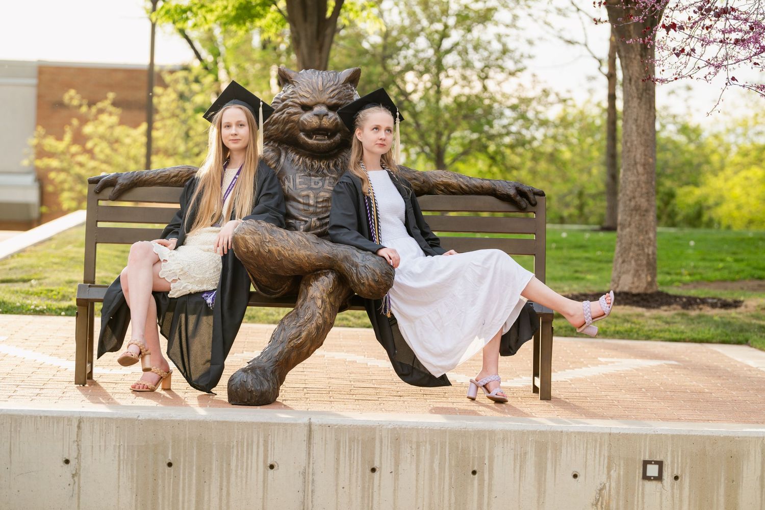 Graduation portrait of two college graduates sitting beside Weber State University mascot statue during spring commencement