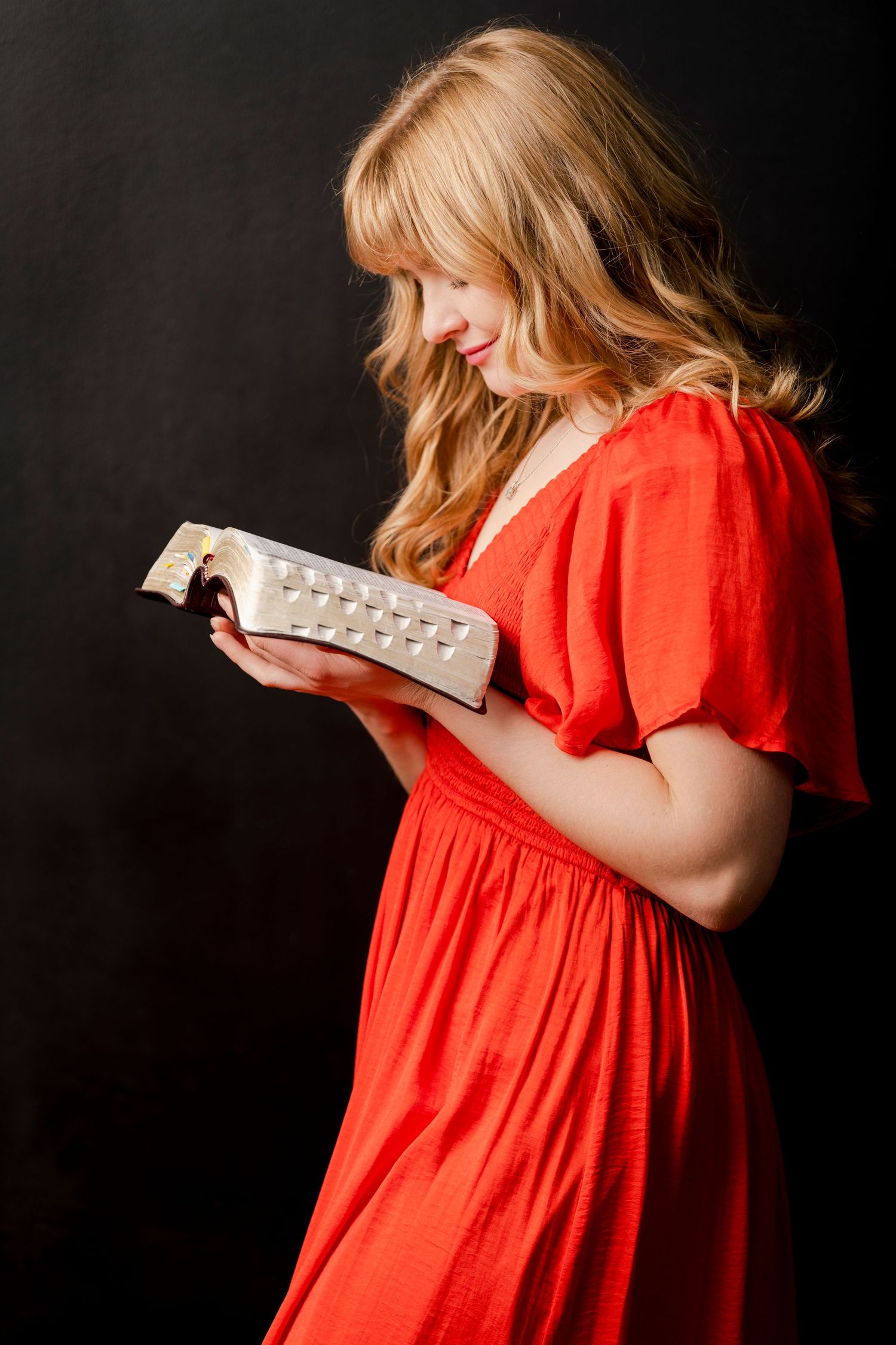 Young missionary woman in red dress reading scriptures in studio with dark background and soft dramatic lighting
