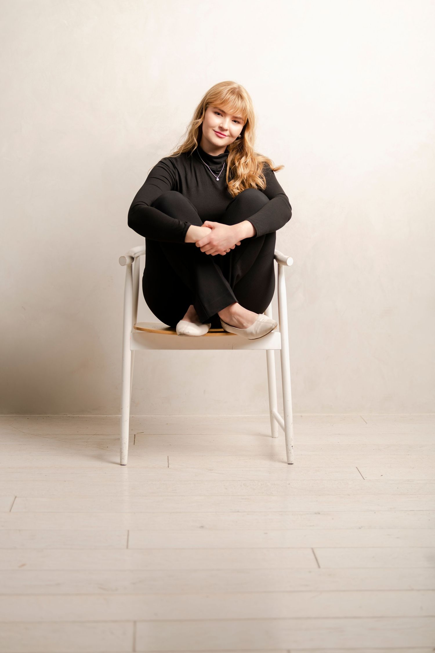 Young woman sitting on chair in studio wearing black outfit with soft light and minimalist background