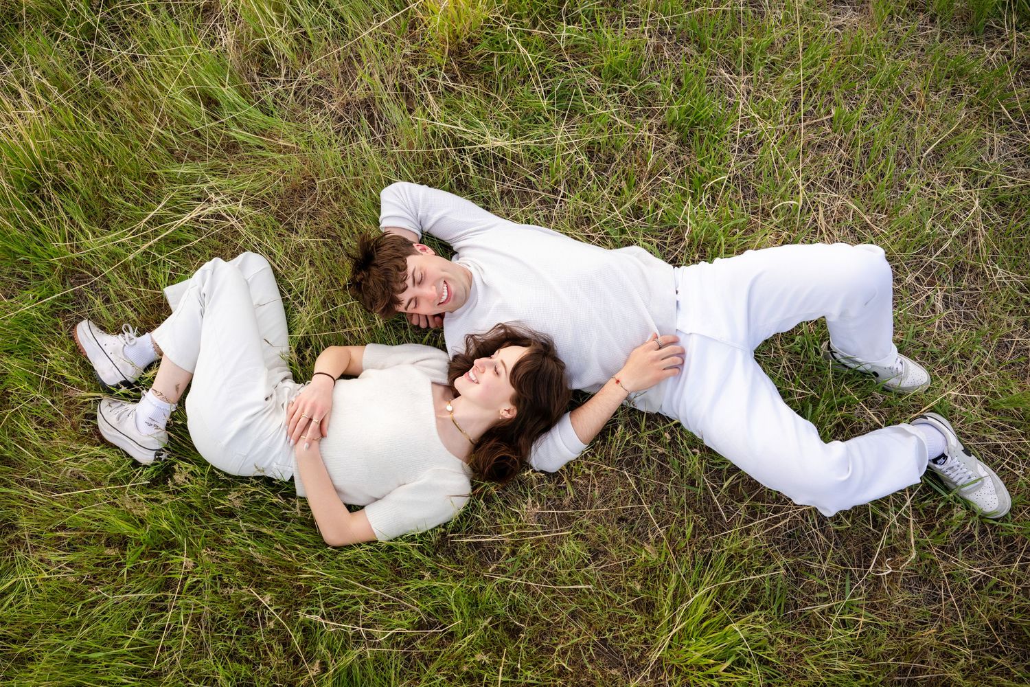 Couple dressed in off-white casually lying down together in the grass in opposite directions looking at each other while smiling and talking