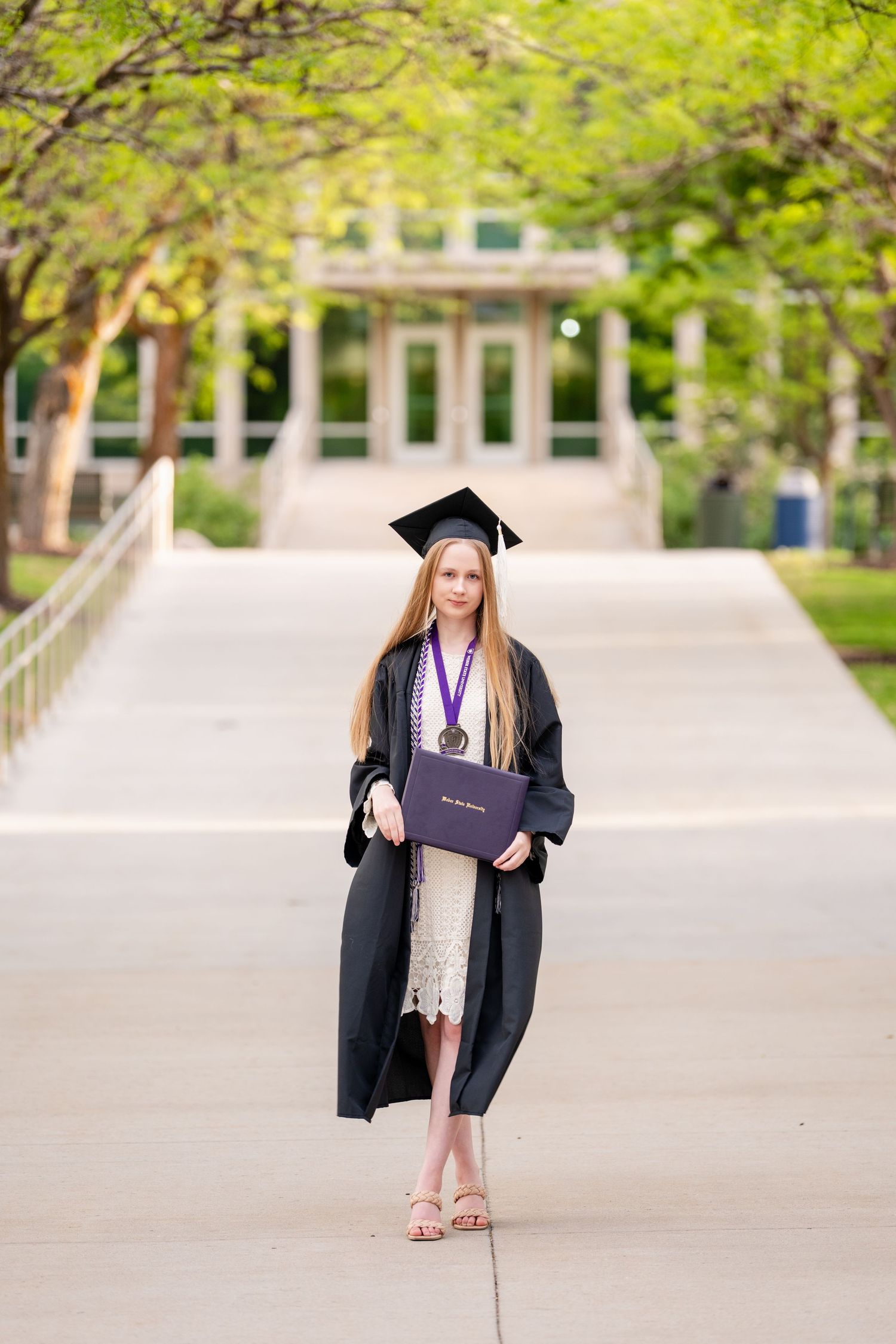 College graduation portrait of female graduate in cap and gown walking through tree lined campus pathway