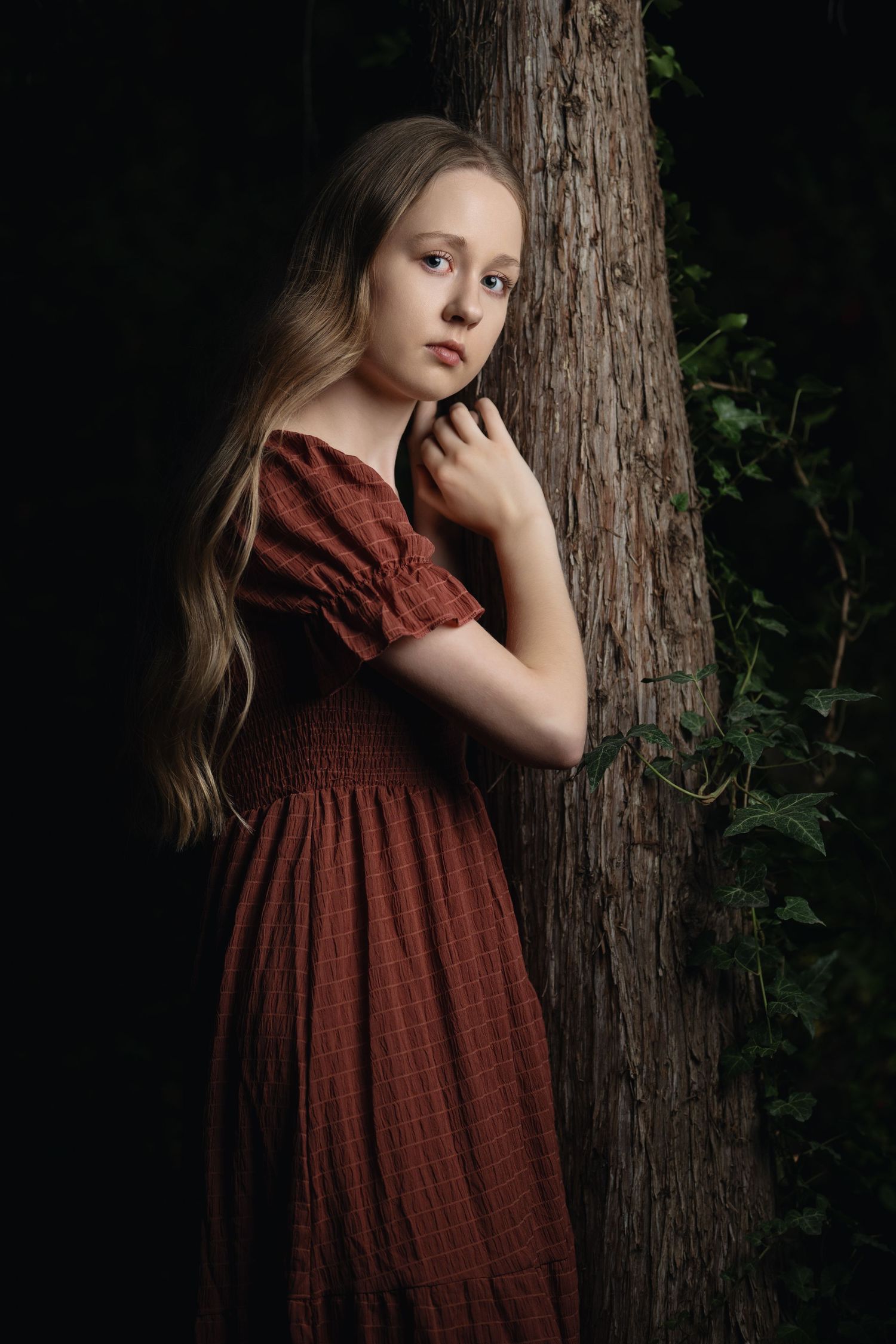 Young woman portrait in red dress standing by tree with dramatic lighting and dark background