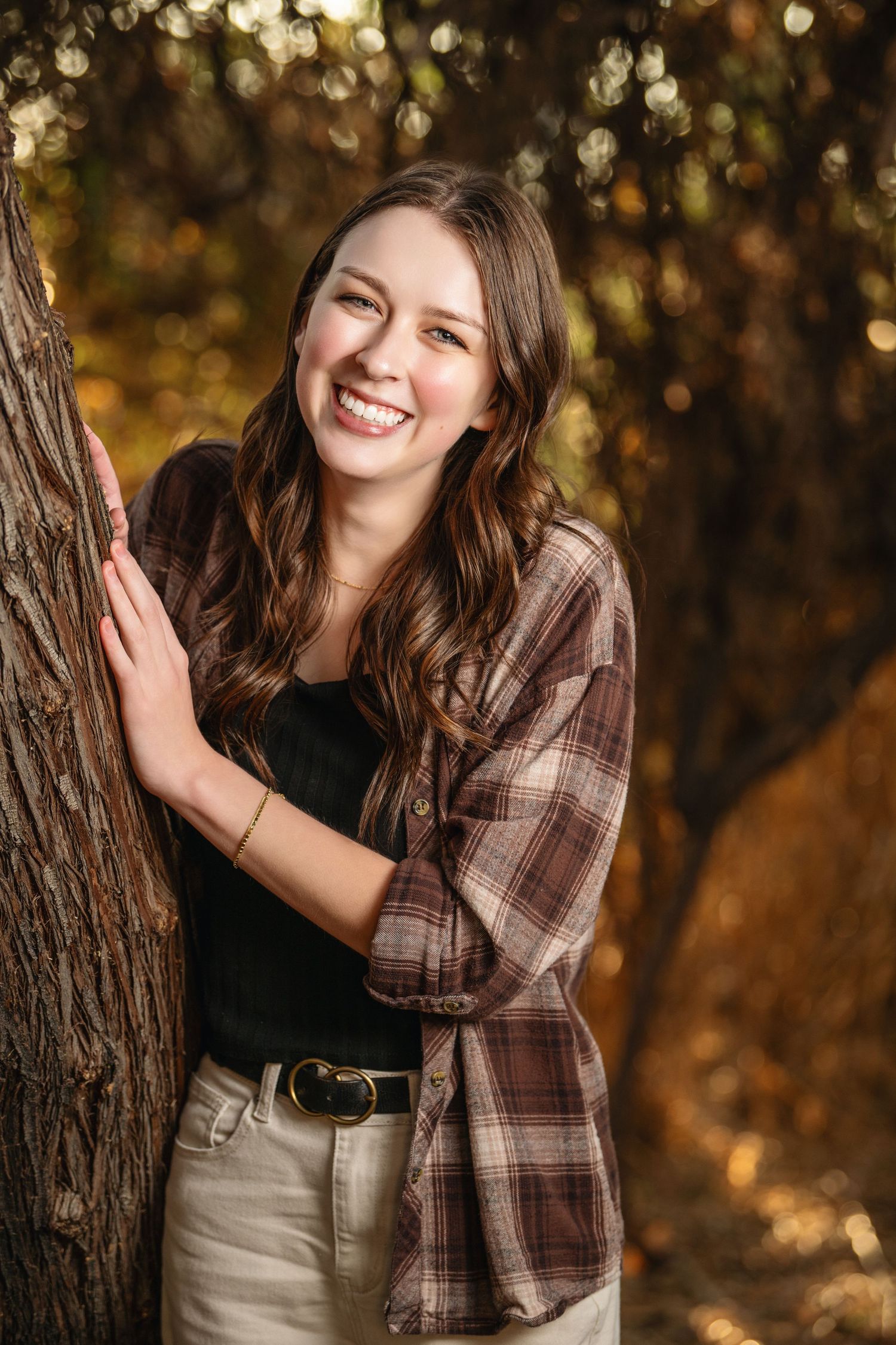 Senior girl smiling by tree in warm autumn light wearing flannel and casual outfit