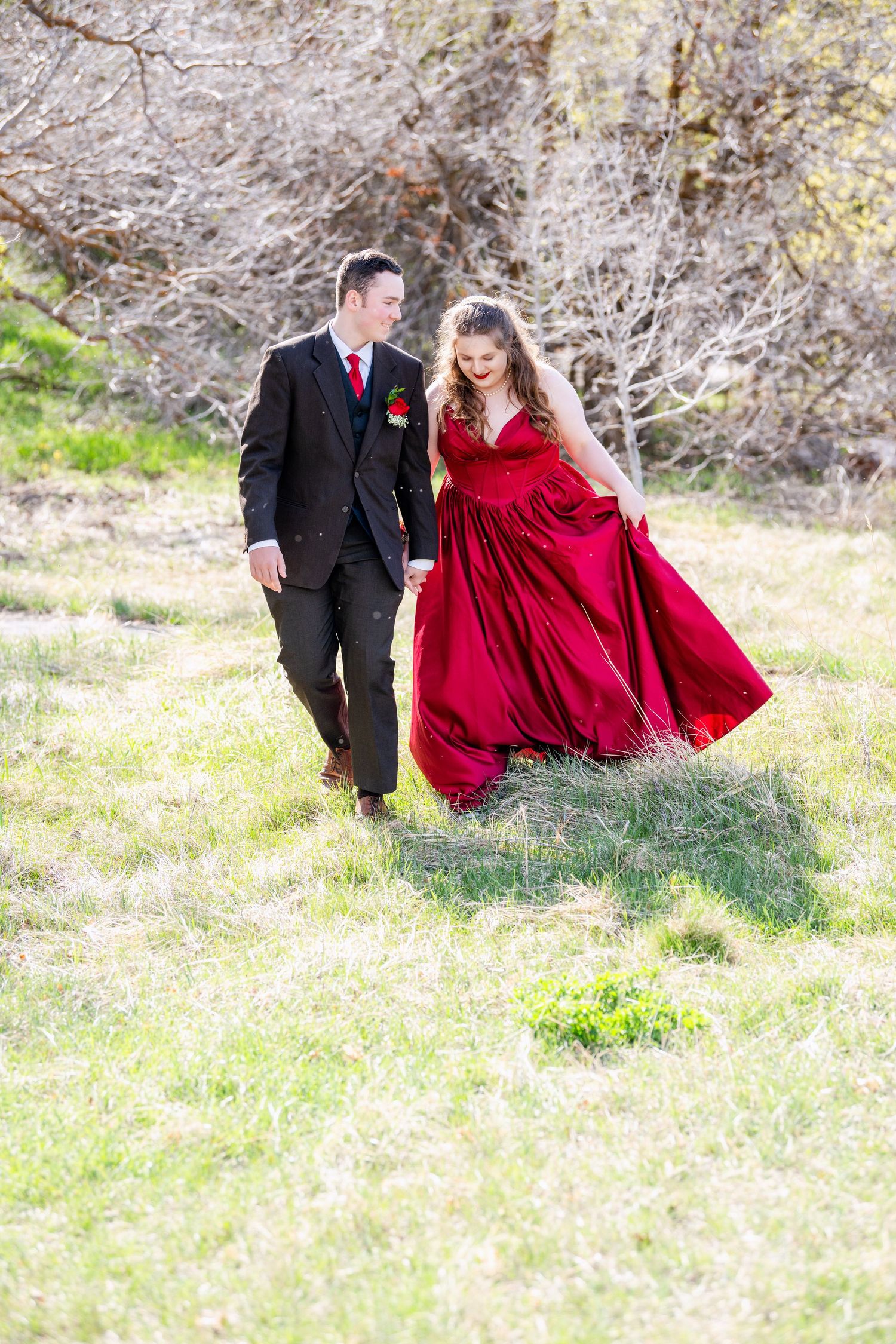 Prom couple walking in grassy field in Woods Cross Utah with flowing red dress and soft natural light