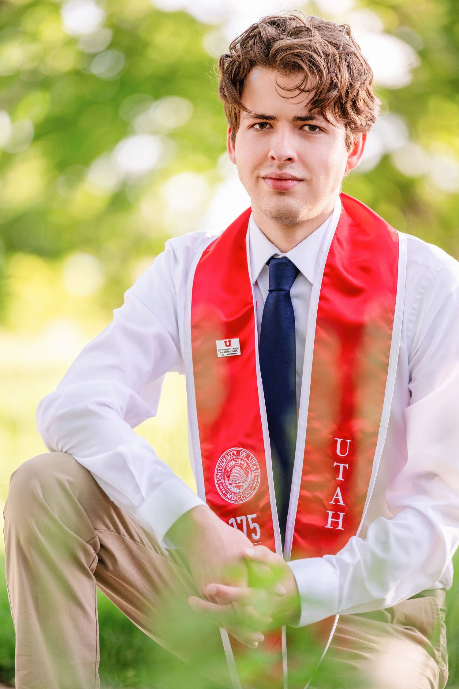 Male college graduate sitting outdoors at the University of Utah campus wearing red stole and tie with bright natural light