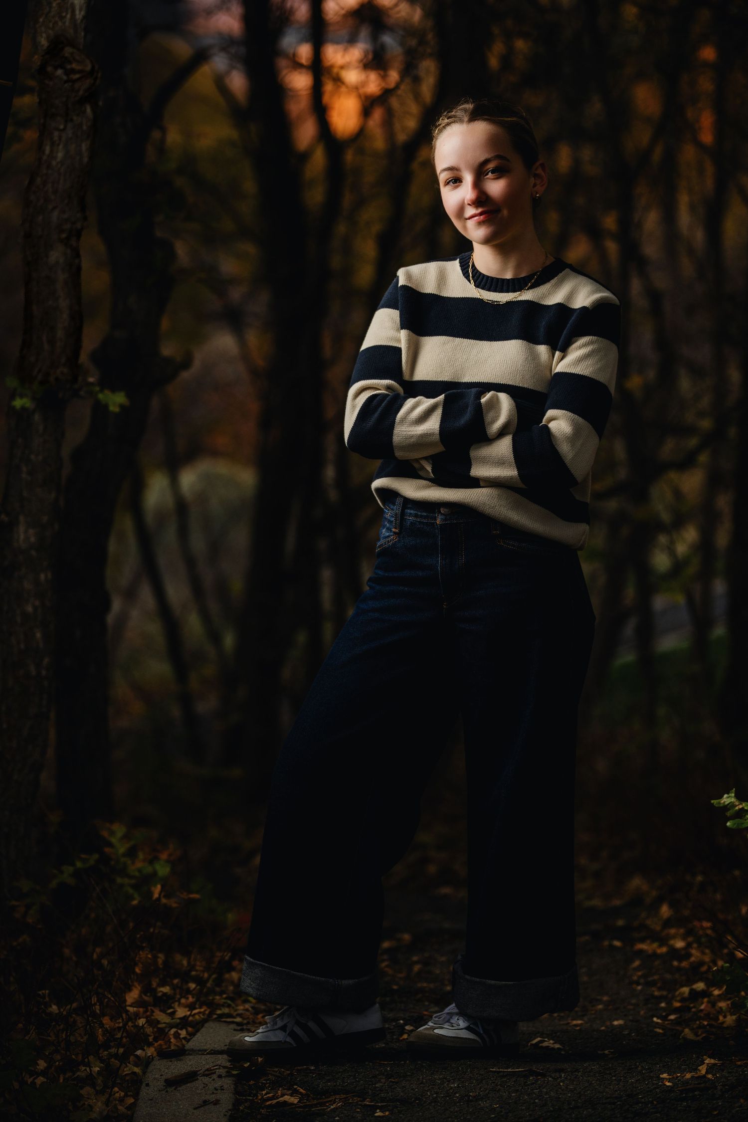 Teen girl standing in North Salt Lake wooded area wearing striped sweater with moody fall tones at sunset