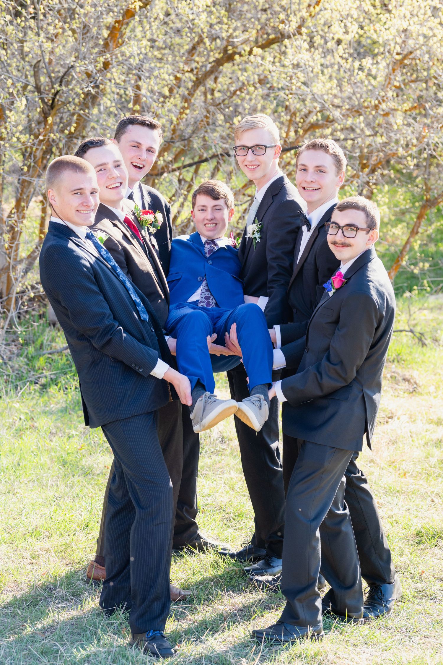 Group of boys in suits lifting friend in blue suit outdoors with playful energy for Woods Cross Prom