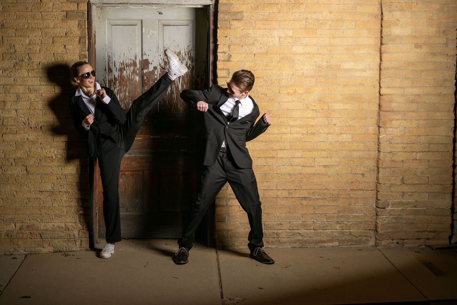 Playful Sadie Hawkins dance portrait with students posing in suits against urban brick wall background