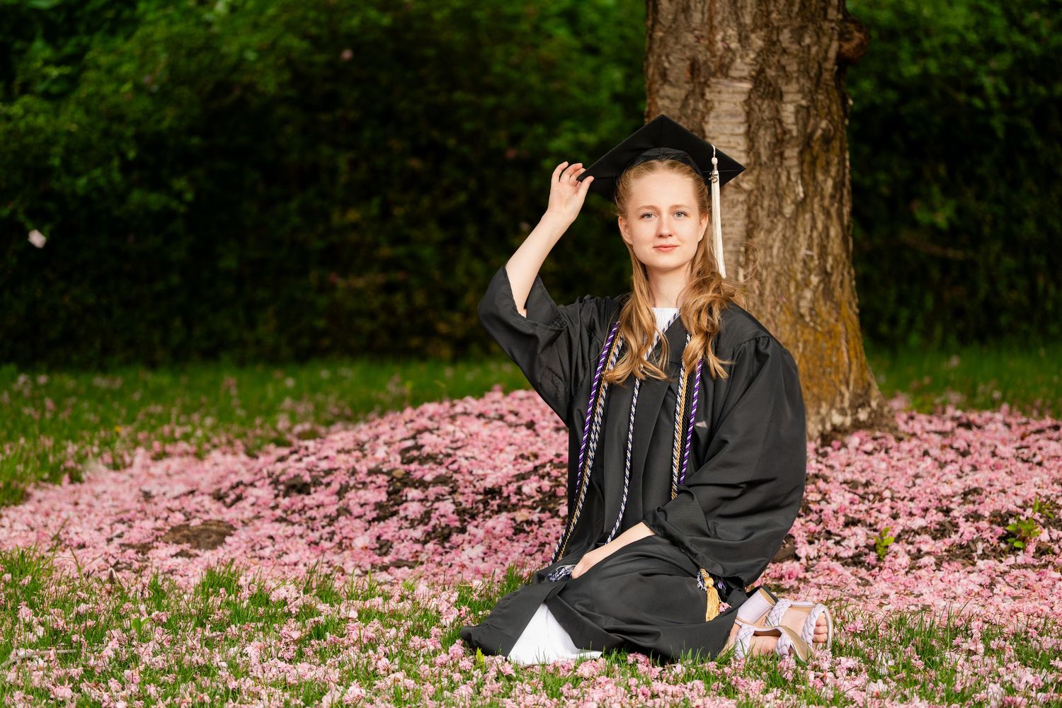 Female graduate sitting in flower petals wearing cap and gown during outdoor college graduation session