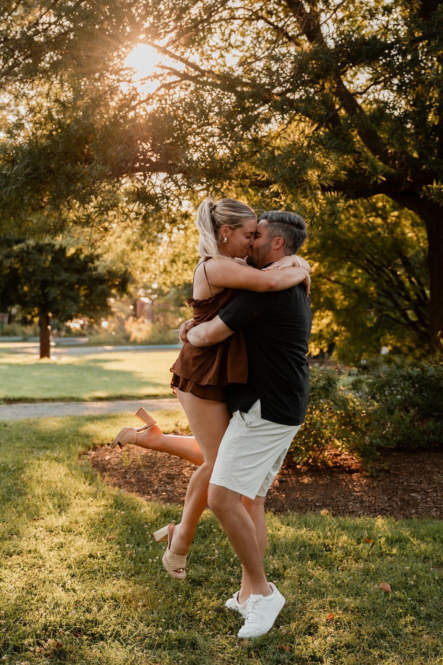 Couple sharing a romantic kiss in a sunlit park, man lifting woman in brown dress amid lush green trees.