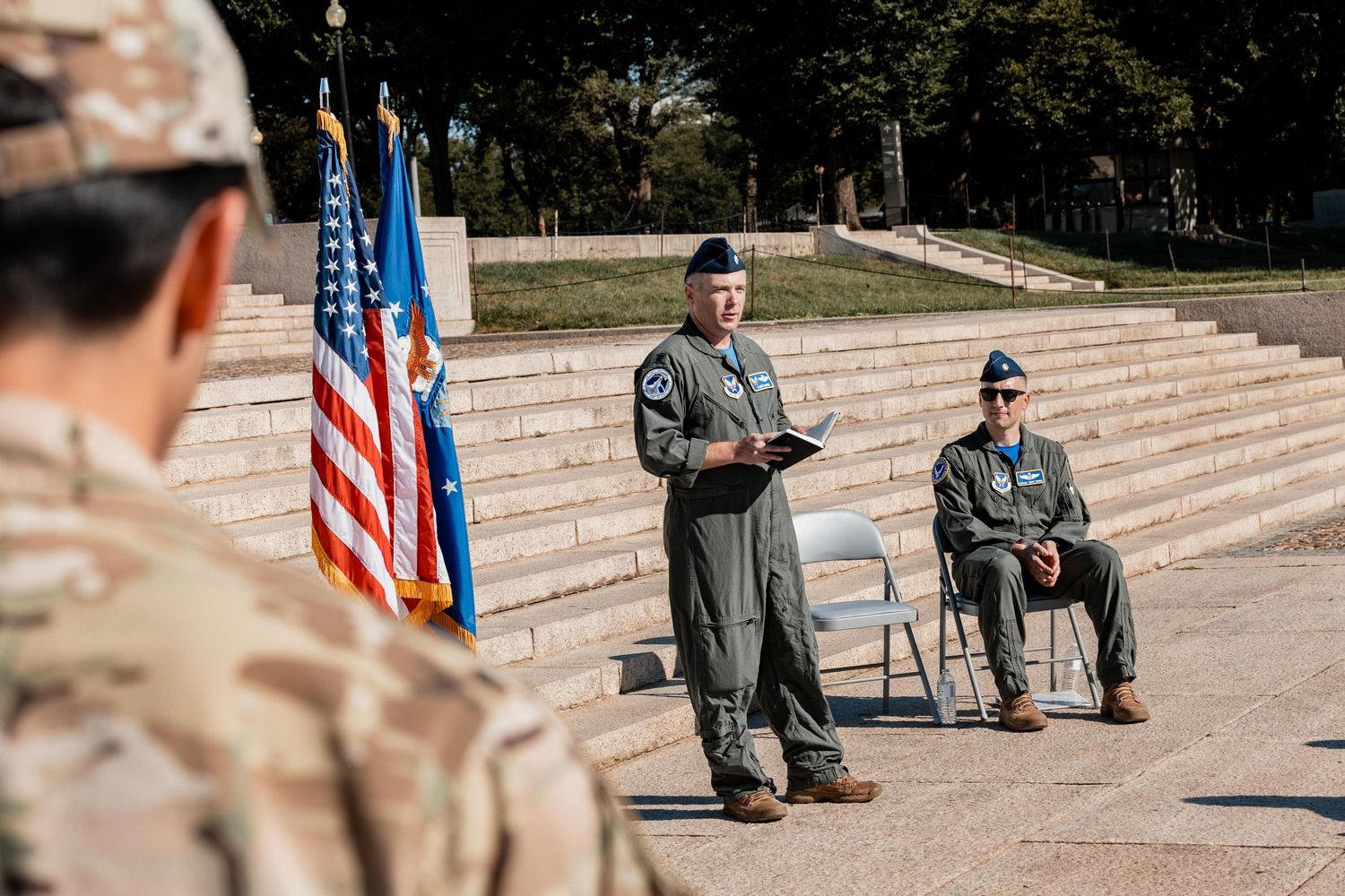 Military officer in flight suit speaks at outdoor ceremony with US and Air Force flags and seated official.