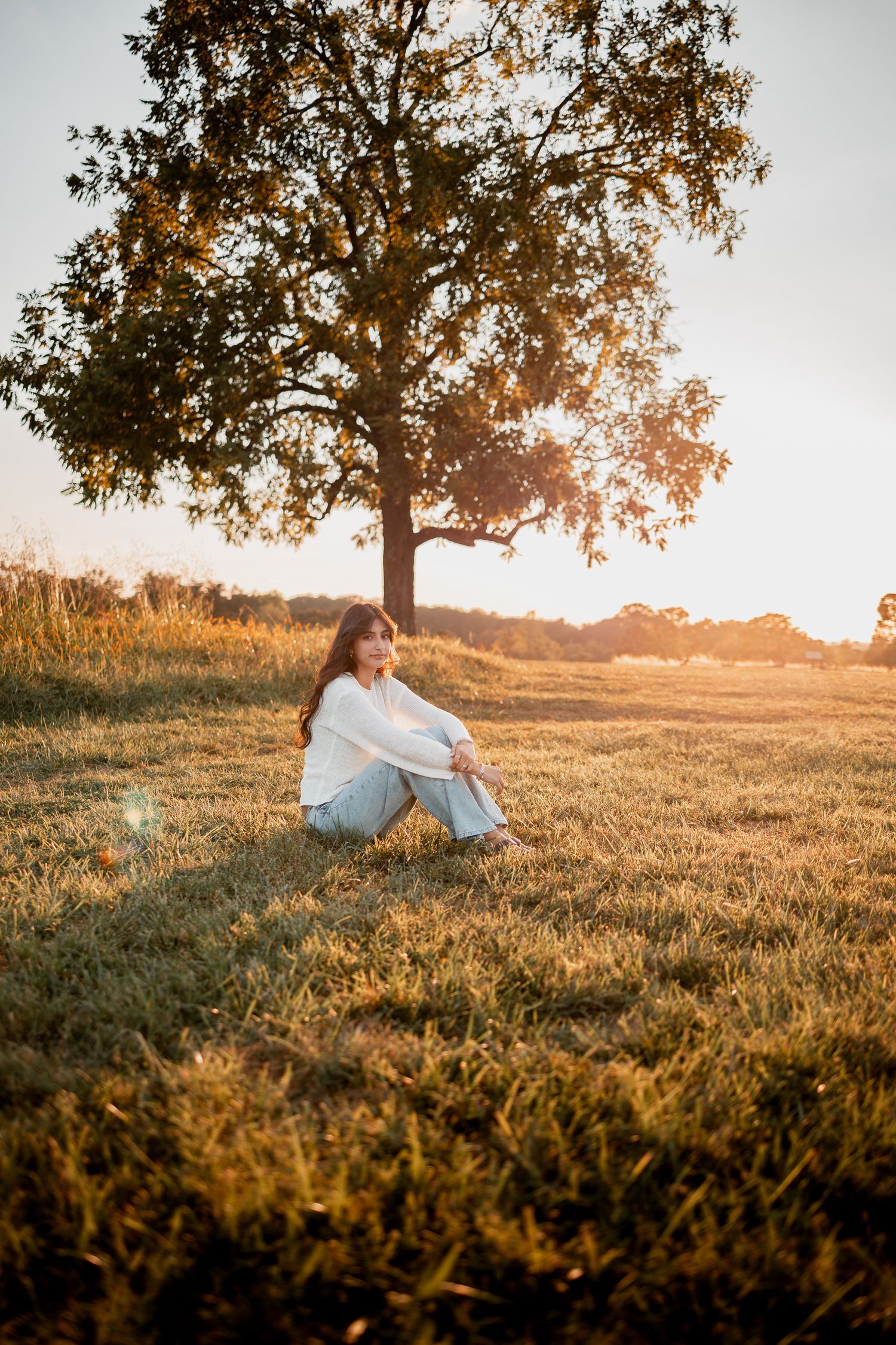 Person sitting in a grassy field at golden hour as sunlight filters through trees in a dreamy outdoor setting.