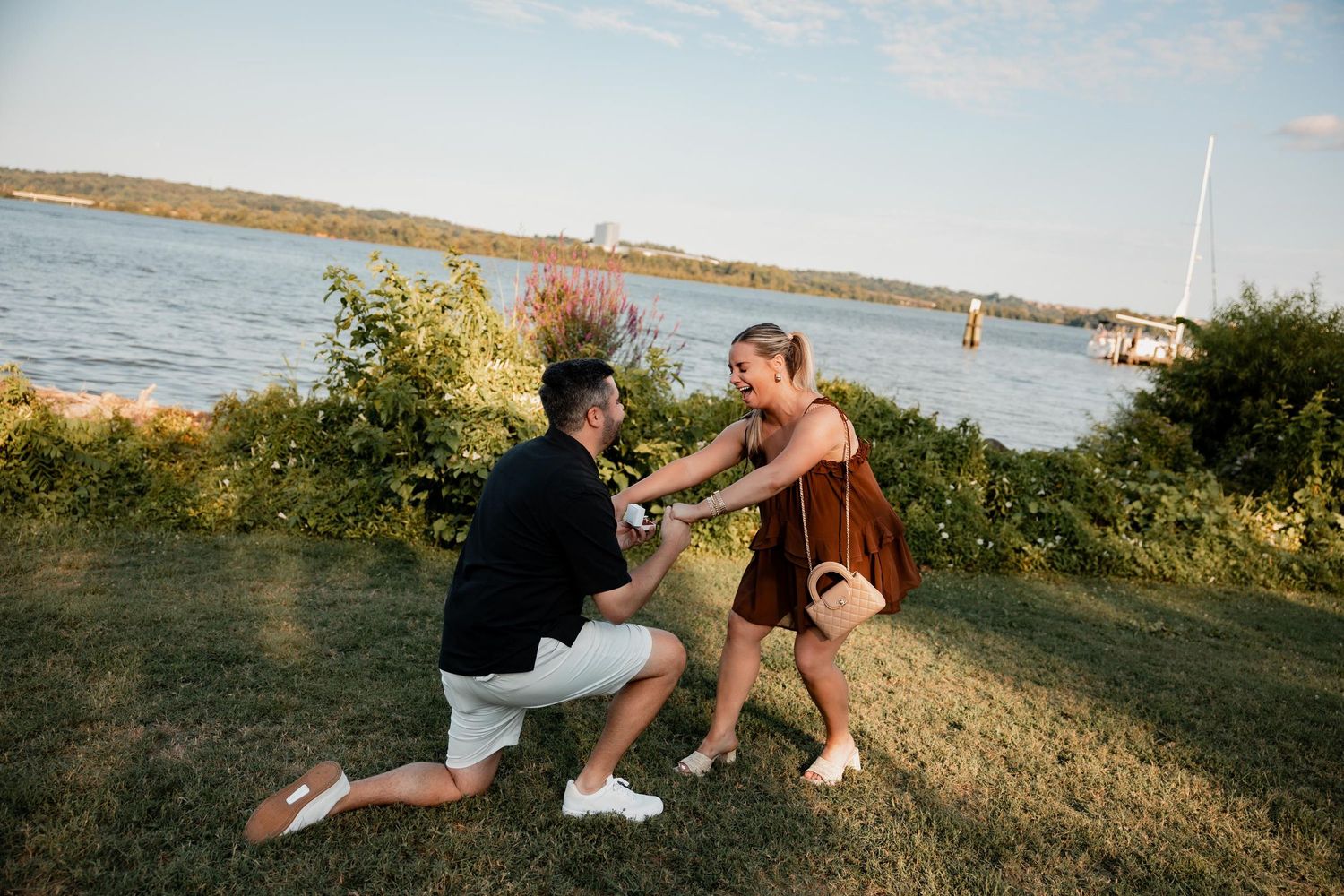Man proposing on one knee to woman near waterfront at golden hour, sailboat visible in background.