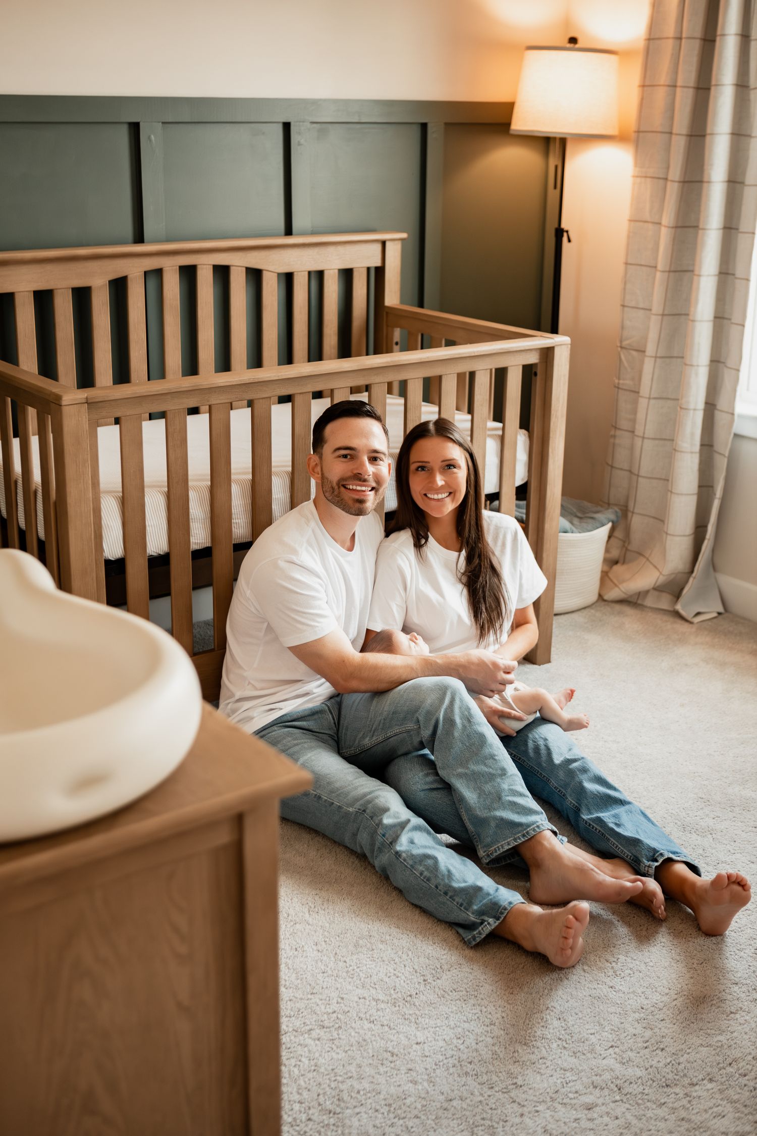 A couple in white shirts and jeans sits casually together on a carpeted floor against a wooden headboard.