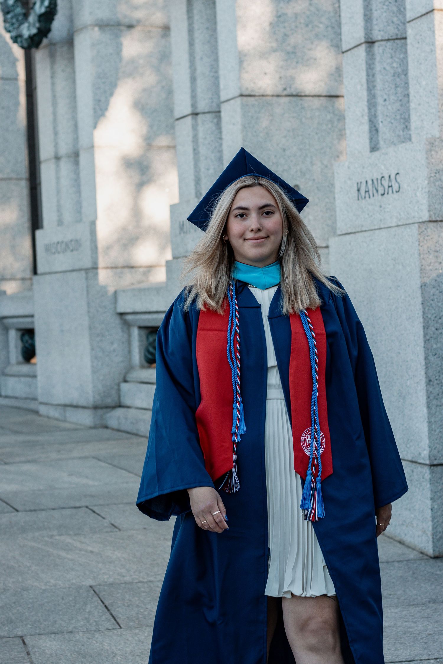 Graduate in navy regalia and red stole stands against stone pillars during a graduation photoshoot.