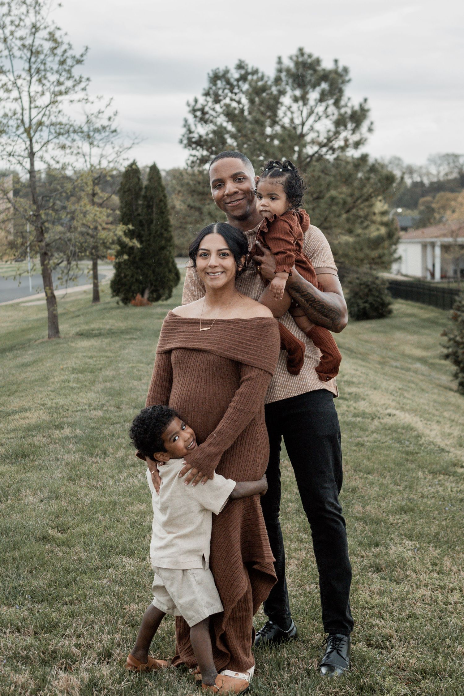 A family wearing coordinating brown and cream outfits poses together on a grassy hillside on an overcast autumn day.