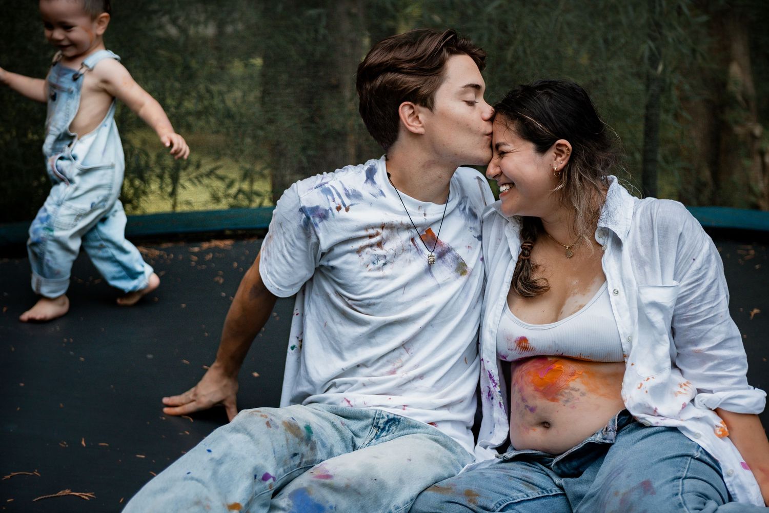A couple in white shirts sits intimately on a trampoline while a child plays in the background.