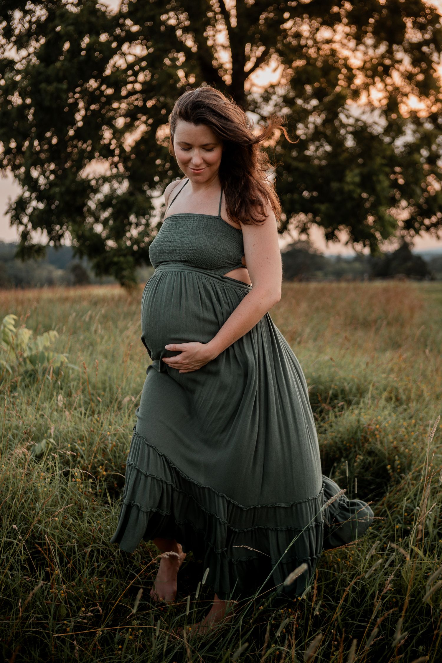 A maternity portrait in a green maxi dress standing in a grassy field at sunset.