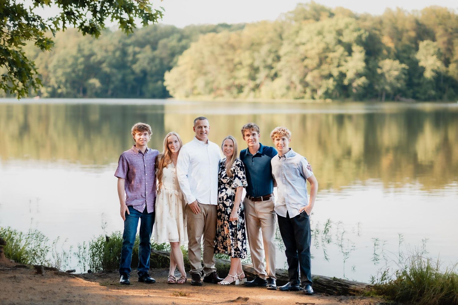 Family of six posing together by a serene lake surrounded by lush green trees during golden hour.