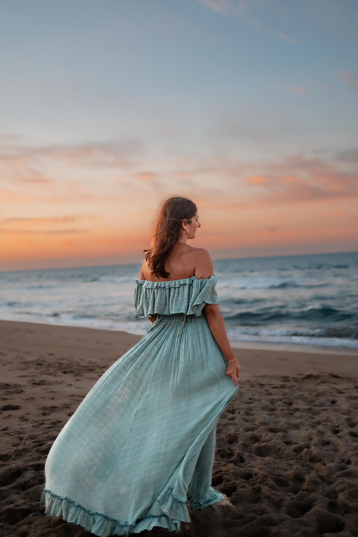 Woman in flowing mint off-shoulder dress stands on sandy beach gazing at vibrant sunset over ocean waves.