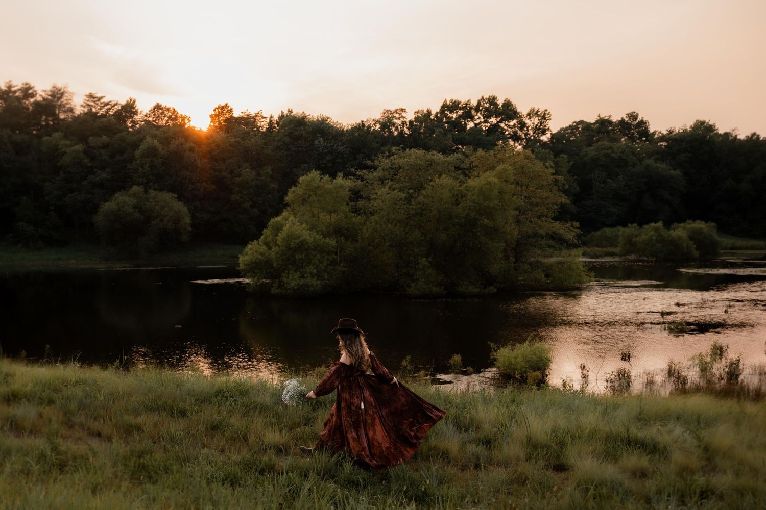 Person in flowing red dress standing by a serene lake at golden sunset with lush green trees reflecting on water.