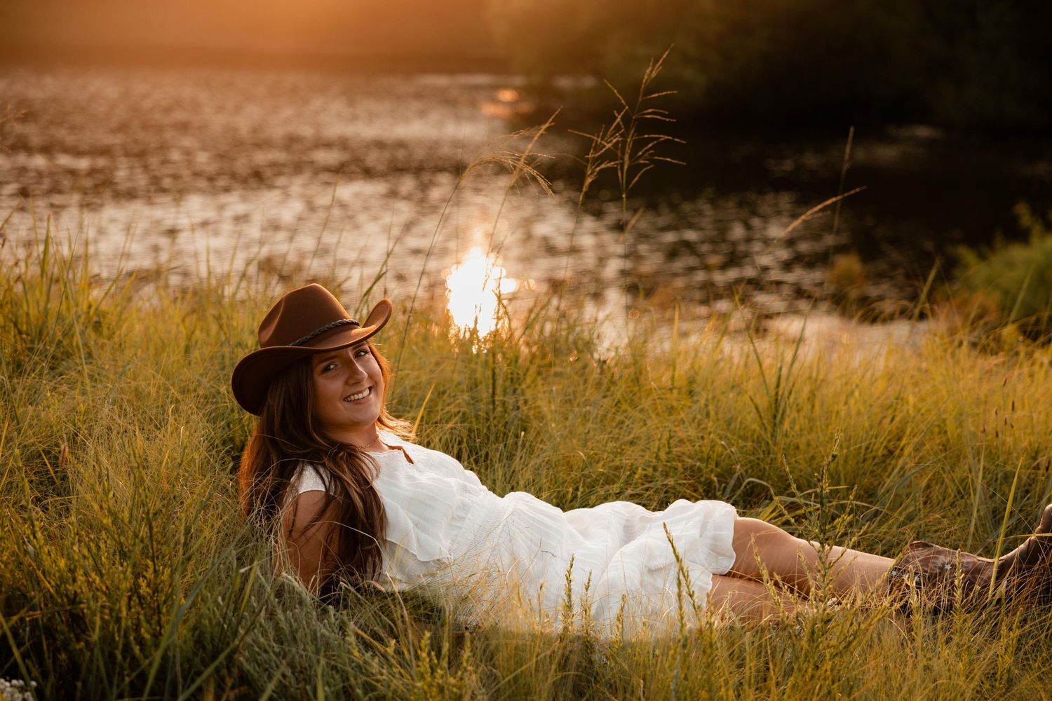 Woman in white dress and cowboy hat relaxing in golden grass by a lake at sunset.