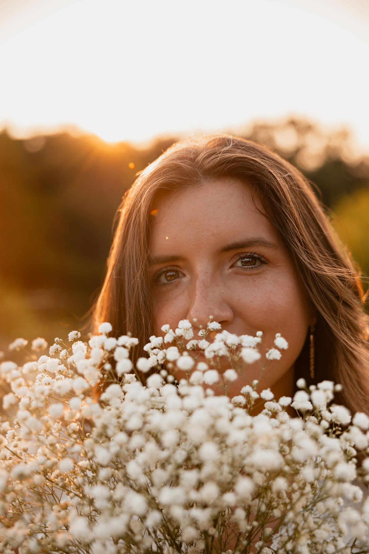 Young woman with freckles holding white baby's breath flowers at golden hour sunset in outdoor field.