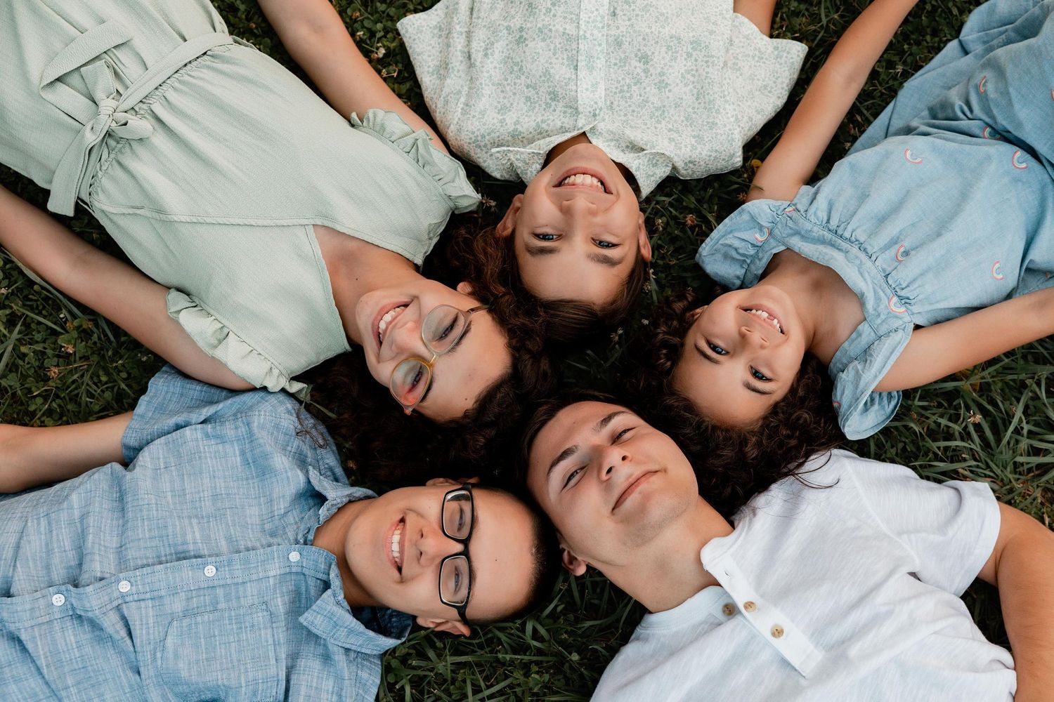 Five young people lying in a circle on grass, smiling and relaxed, viewed from above in casual summer outfits.