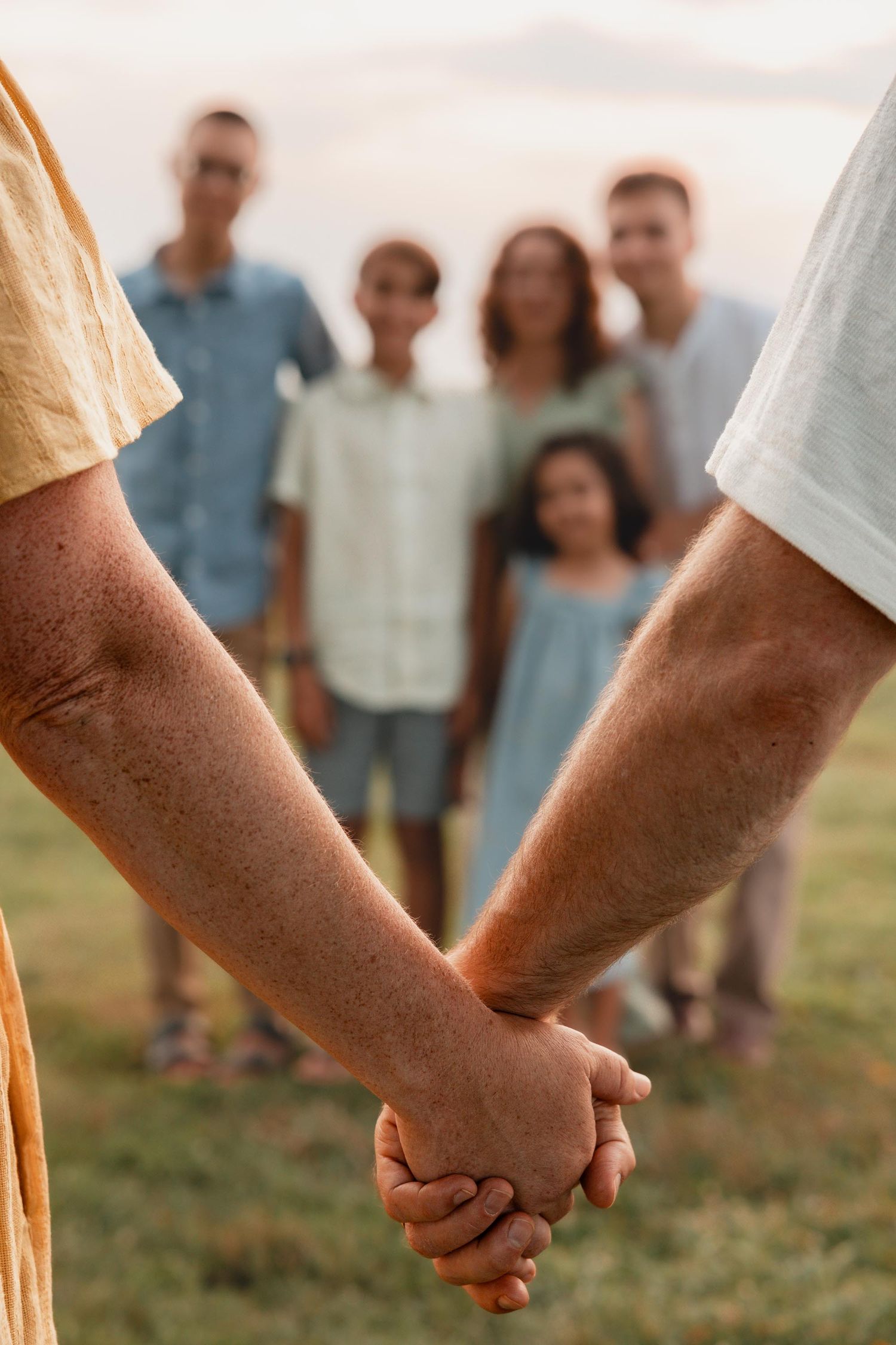 A couple holds hands in the foreground while their three children stand together in a green field.