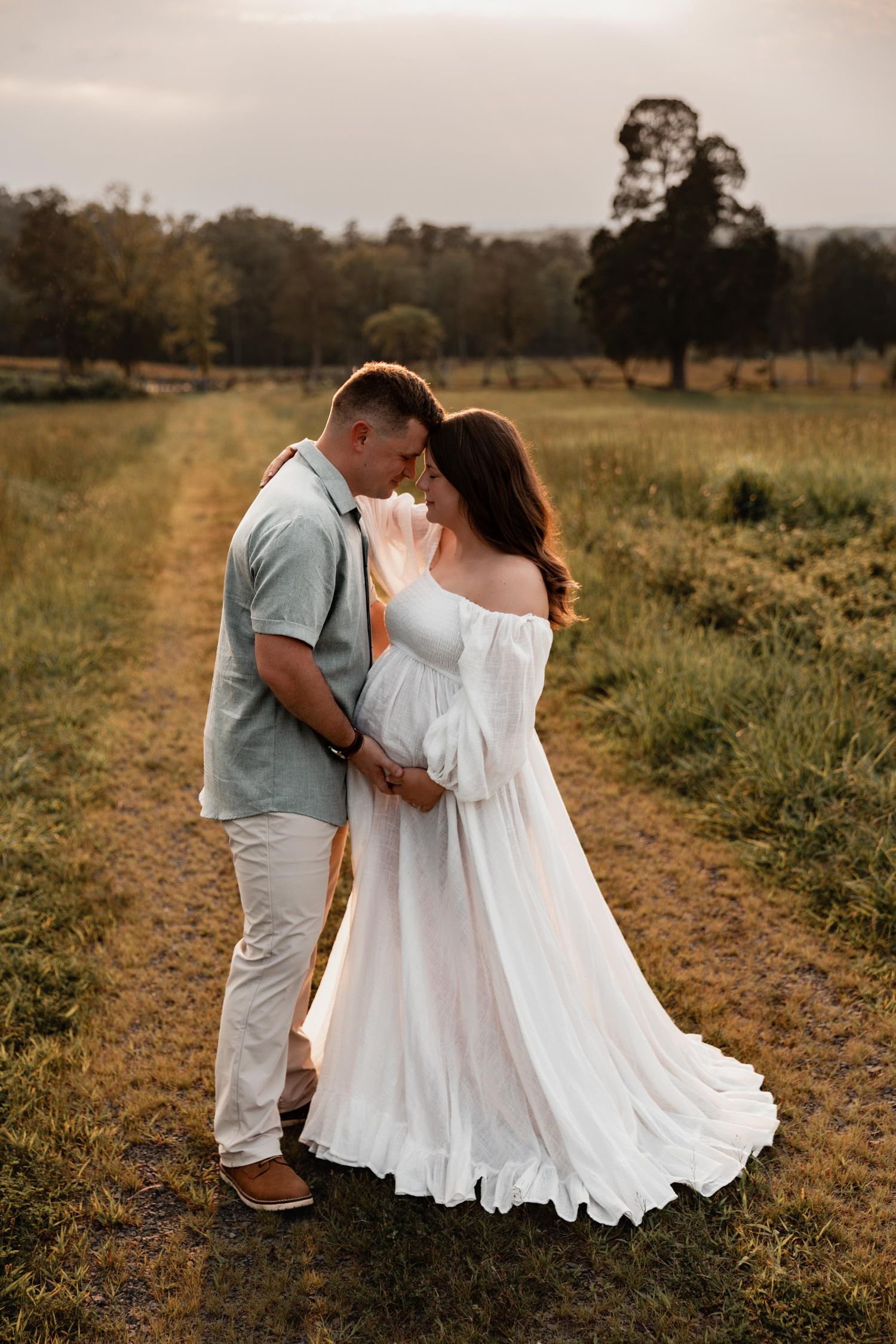 Couple sharing a tender moment on a grassy field path, woman in flowing white maternity gown at golden hour.