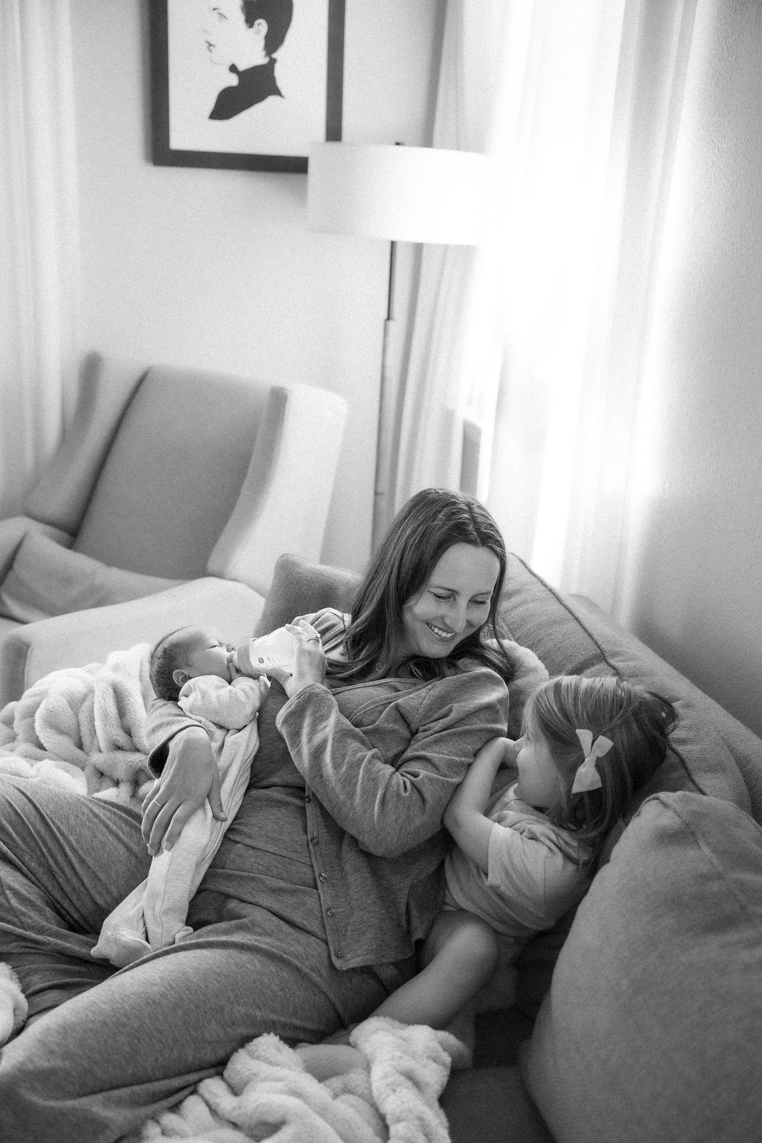 Black and white photo of a smiling mother relaxing on a couch with her newborn baby and young daughter.