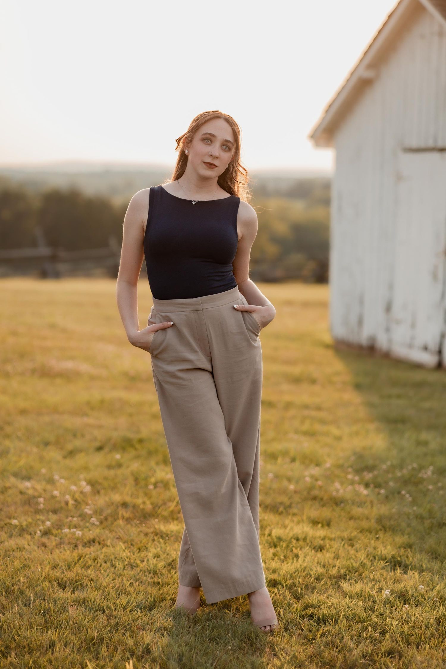 Young woman in black tank top and beige wide-leg pants poses in golden hour sunlight near a white barn.