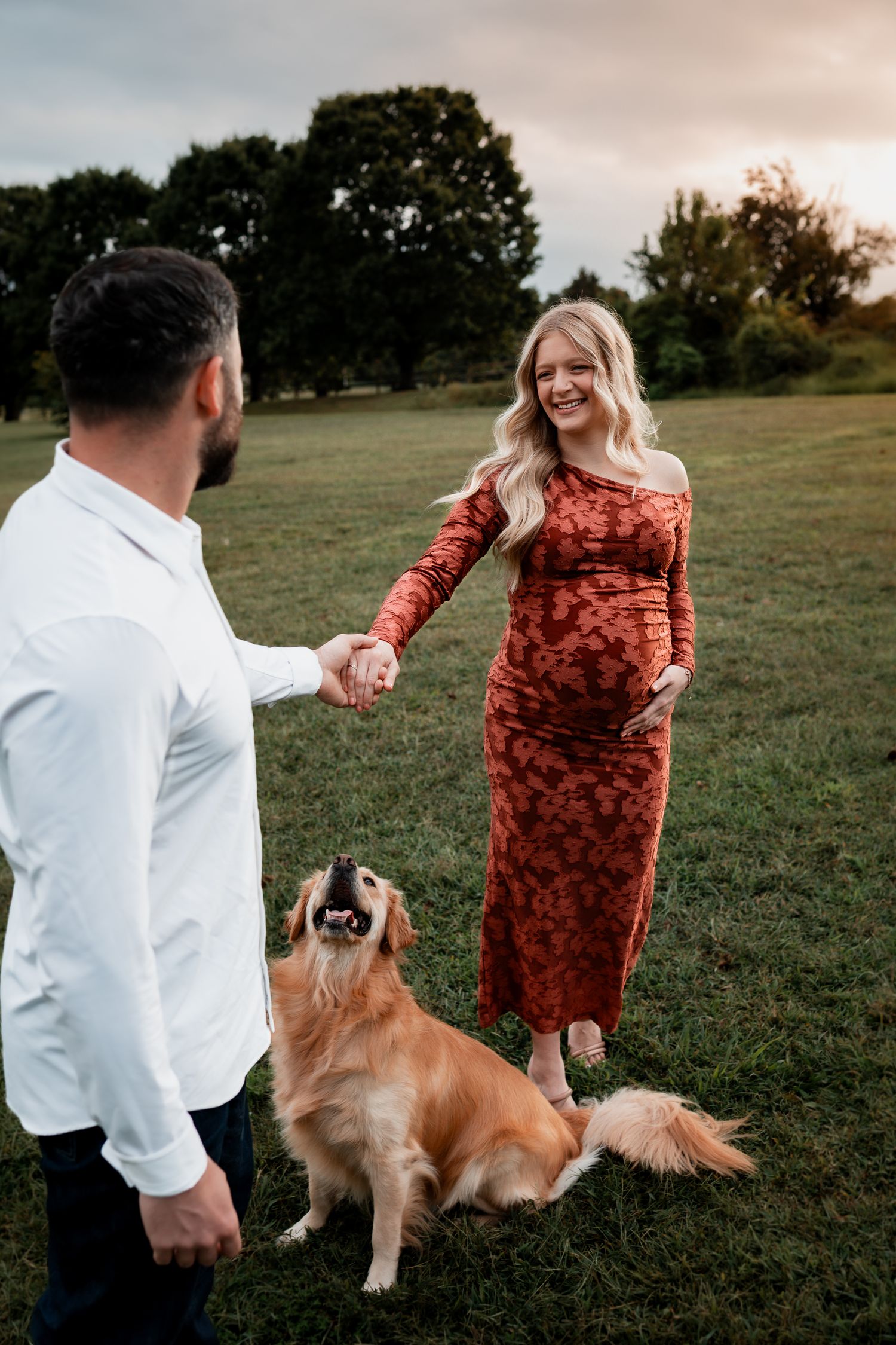 A couple dances with their golden retriever in a scenic outdoor field.